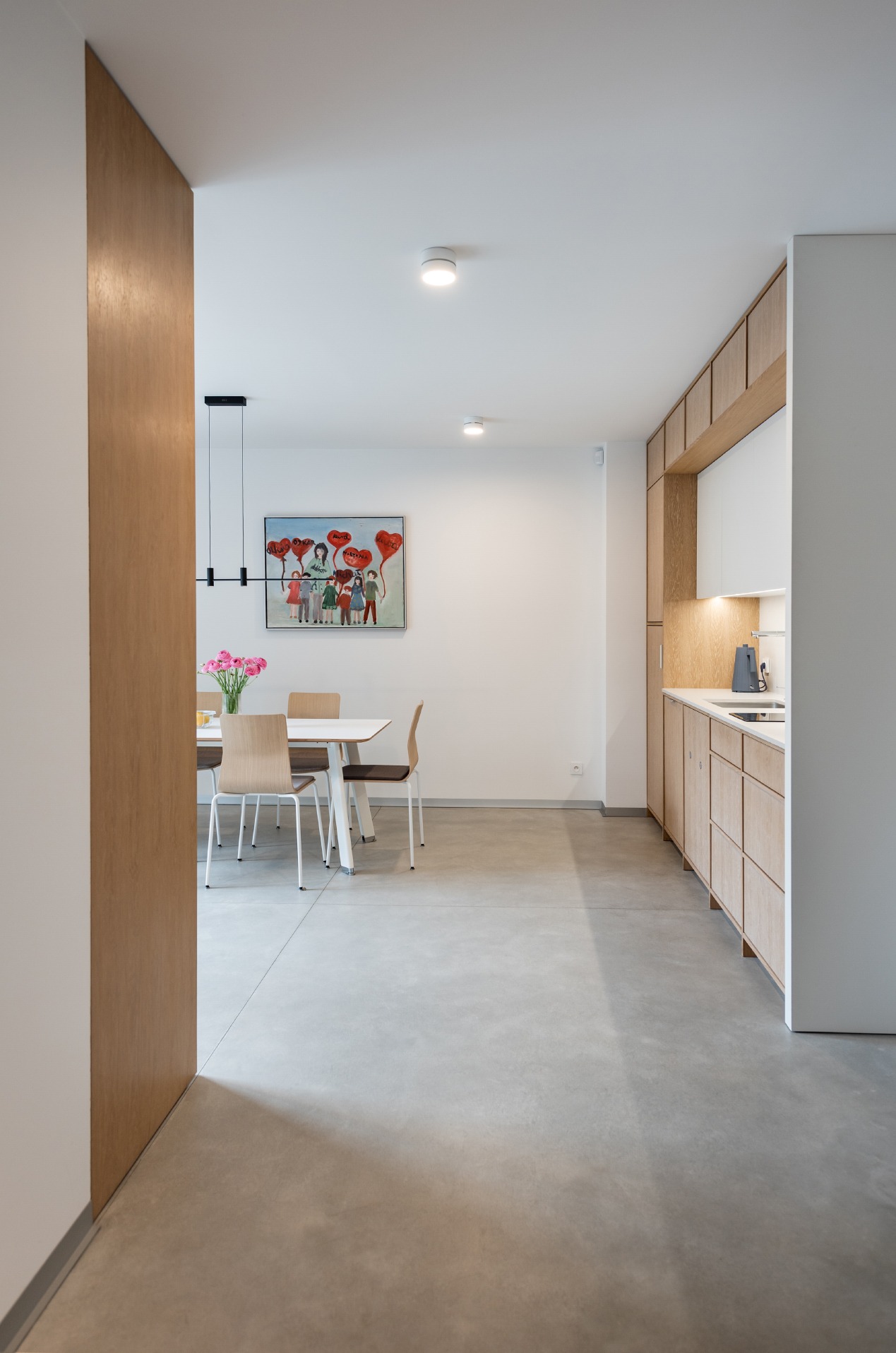 Wnętrze mieszkania w minimalistycznym stylu, z widokiem na jadalnię z białym stołem i drewnianymi krzesłami oraz kuchnię z drewnianymi szafkami, betonowa posadzka, obraz na ścianie.