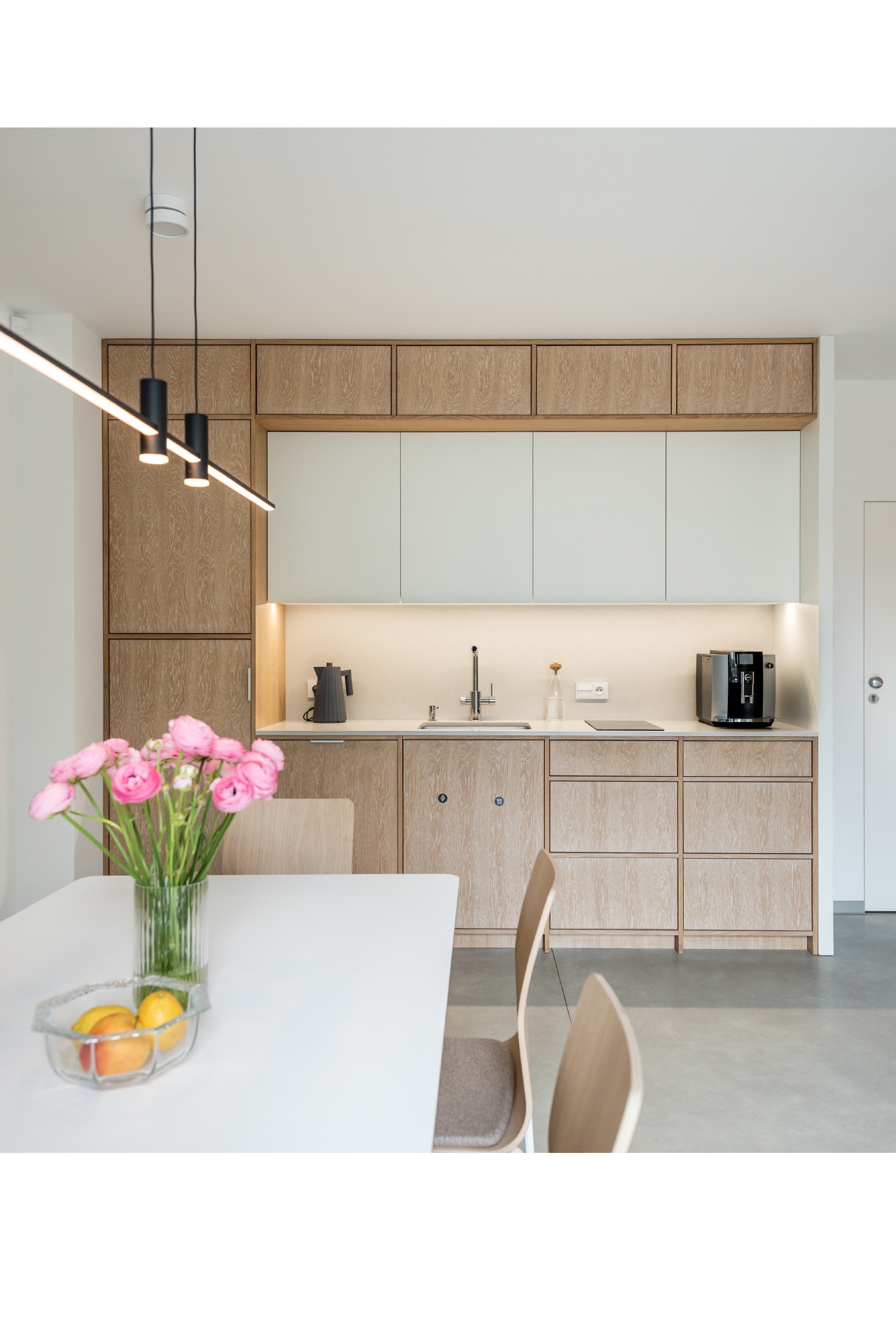Nowoczesna kuchnia z drewnianymi i białymi szafkami, oświetleniem LED pod szafkami górnymi, czarnym ekspresem do kawy i czajnikiem na blacie, oraz wazonem z różowymi kwiatami na białym stole jadalnym.