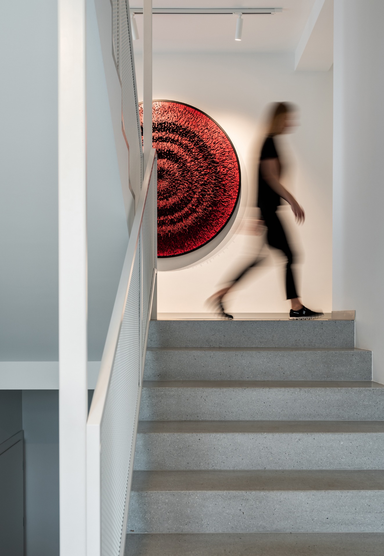 Betonowe schody z białą balustradą, czerwona okrągła dekoracja ścienna, rozmyta sylwetka kobiety schodzącej po schodach.