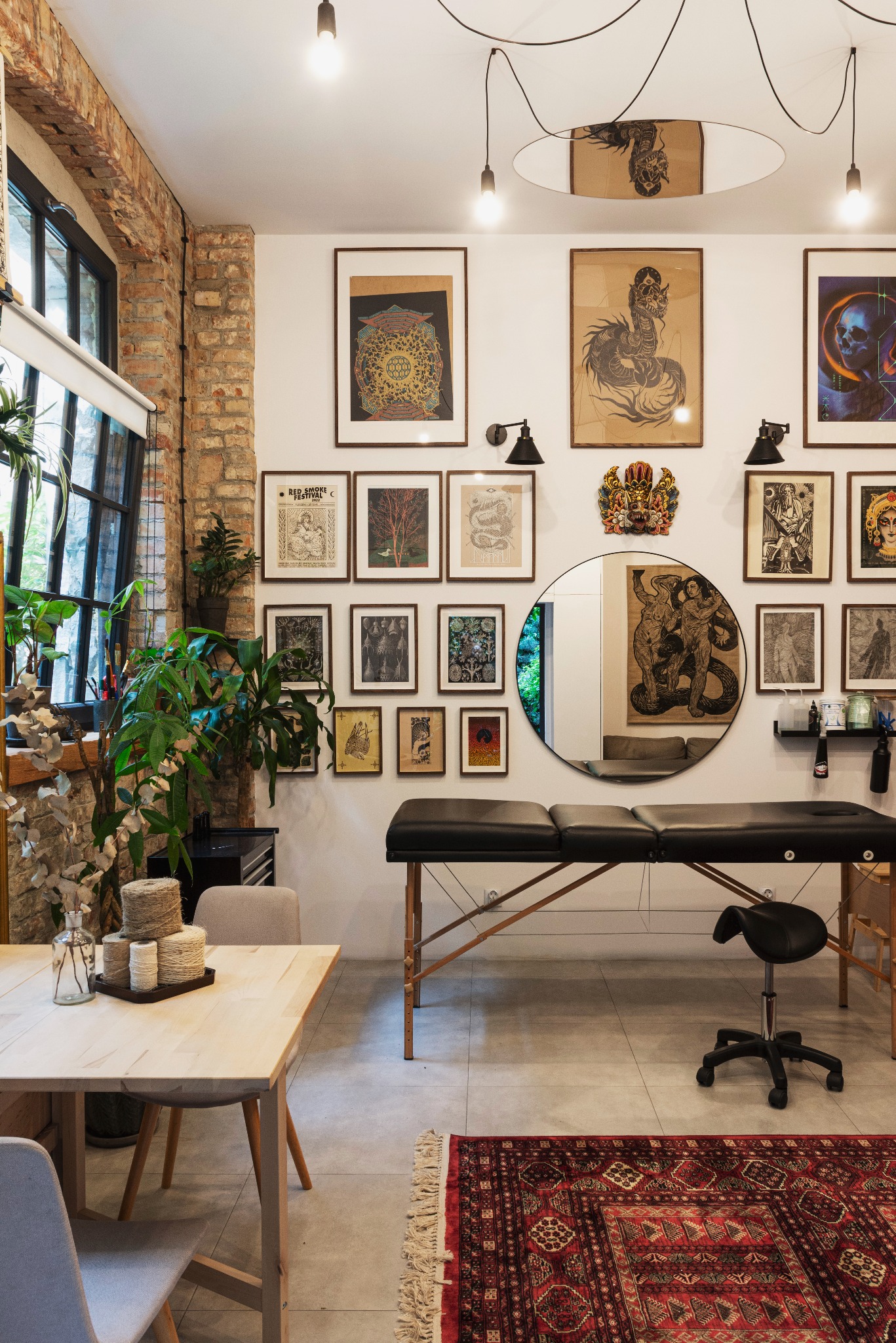 Wnętrze studia z kozetką do zabiegów, ściana udekorowana grafikami w ramkach, okrągłe lustro, drewniany stół z dekoracjami i orientalny dywan.