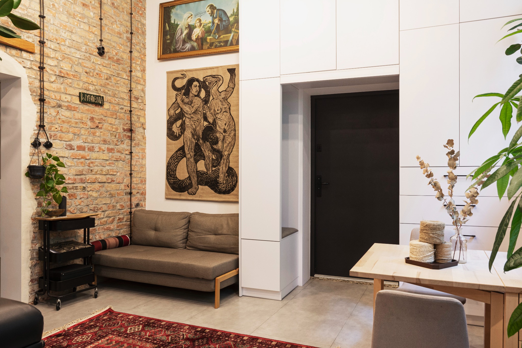 Salon w stylu loft z ceglaną ścianą, szarą sofą, dywanem w orientalne wzory i grafiką na ścianie, widok na stół z dekoracjami.