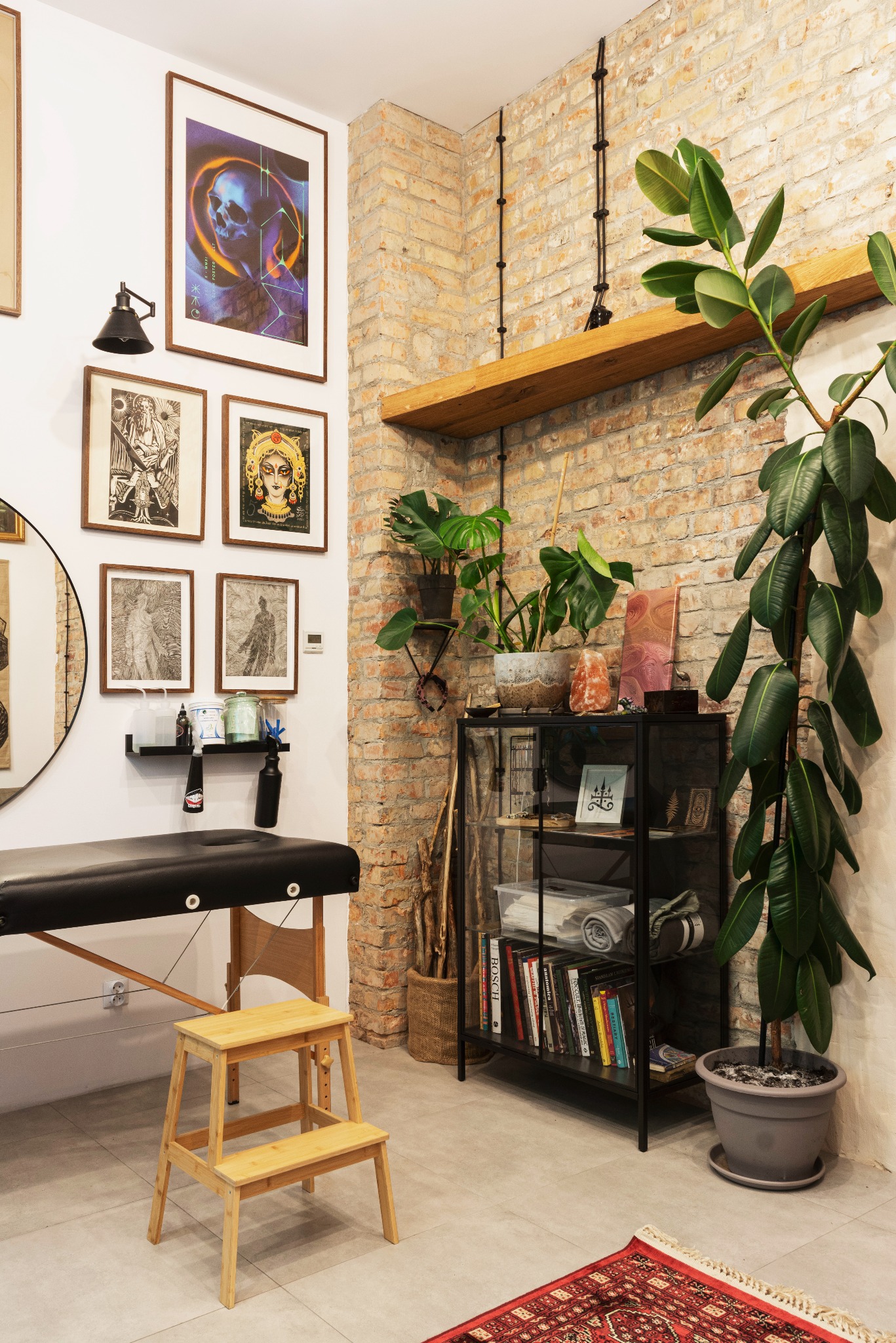 Wnętrze salonu tatuażu z czarnym leżakiem, drewnianym stołkiem schodkowym, regałem z książkami i roślinami, ceglaną ścianą z półką i wiszącymi obrazami.