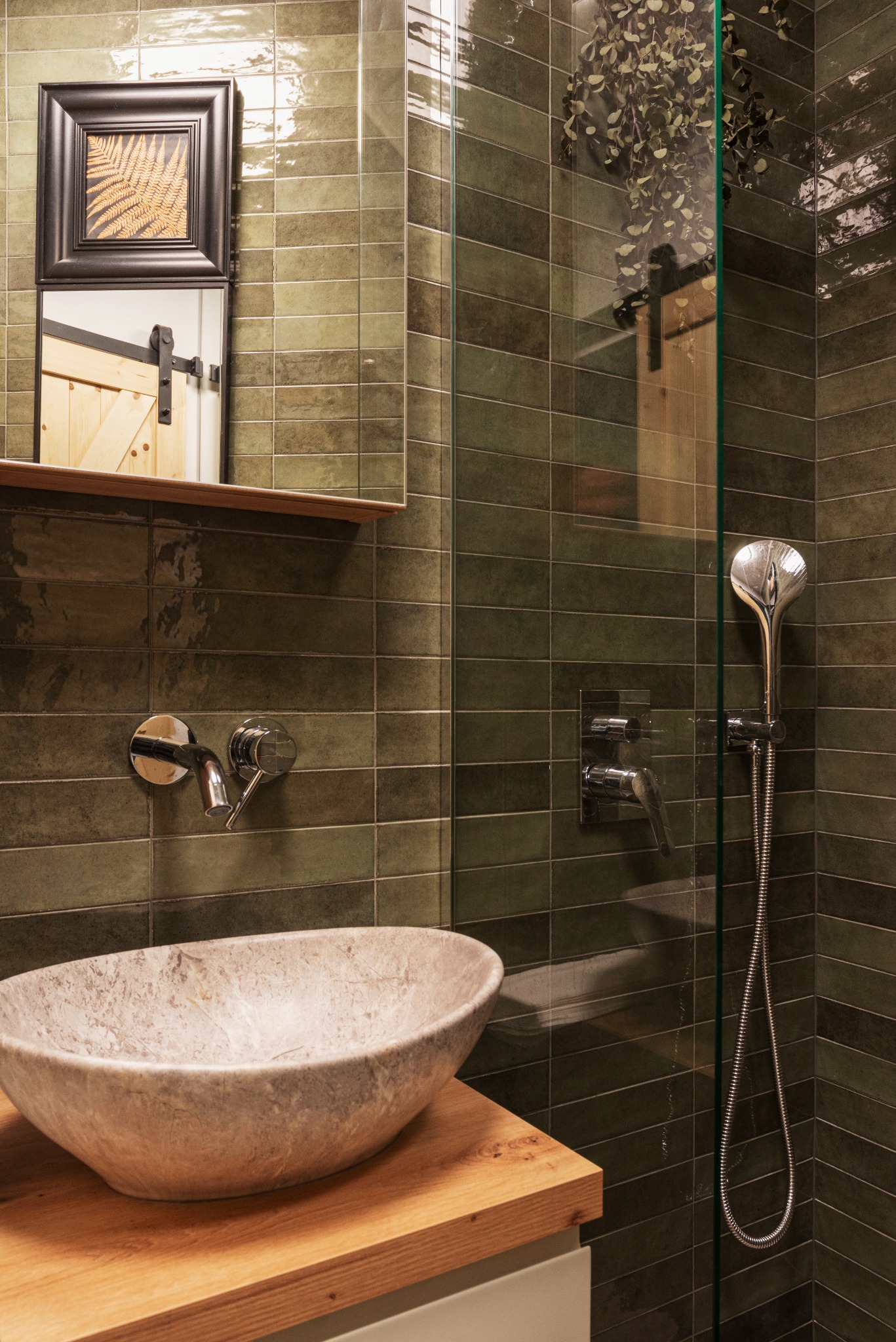 Nowoczesna łazienka z umywalką z kamienia, zielonymi płytkami i kabiną prysznicową ze szklaną ścianką, odbicie drewnianych drzwi w szkle.