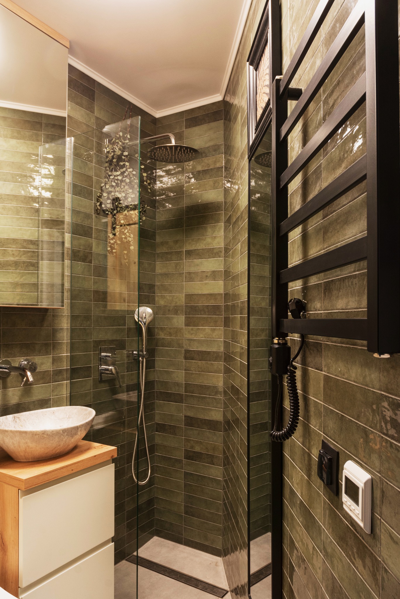 Nowoczesna łazienka z zielonymi, poziomymi płytkami, szklaną kabiną prysznicową, kamienną umywalką nablatową i czarnym grzejnikiem drabinkowym.