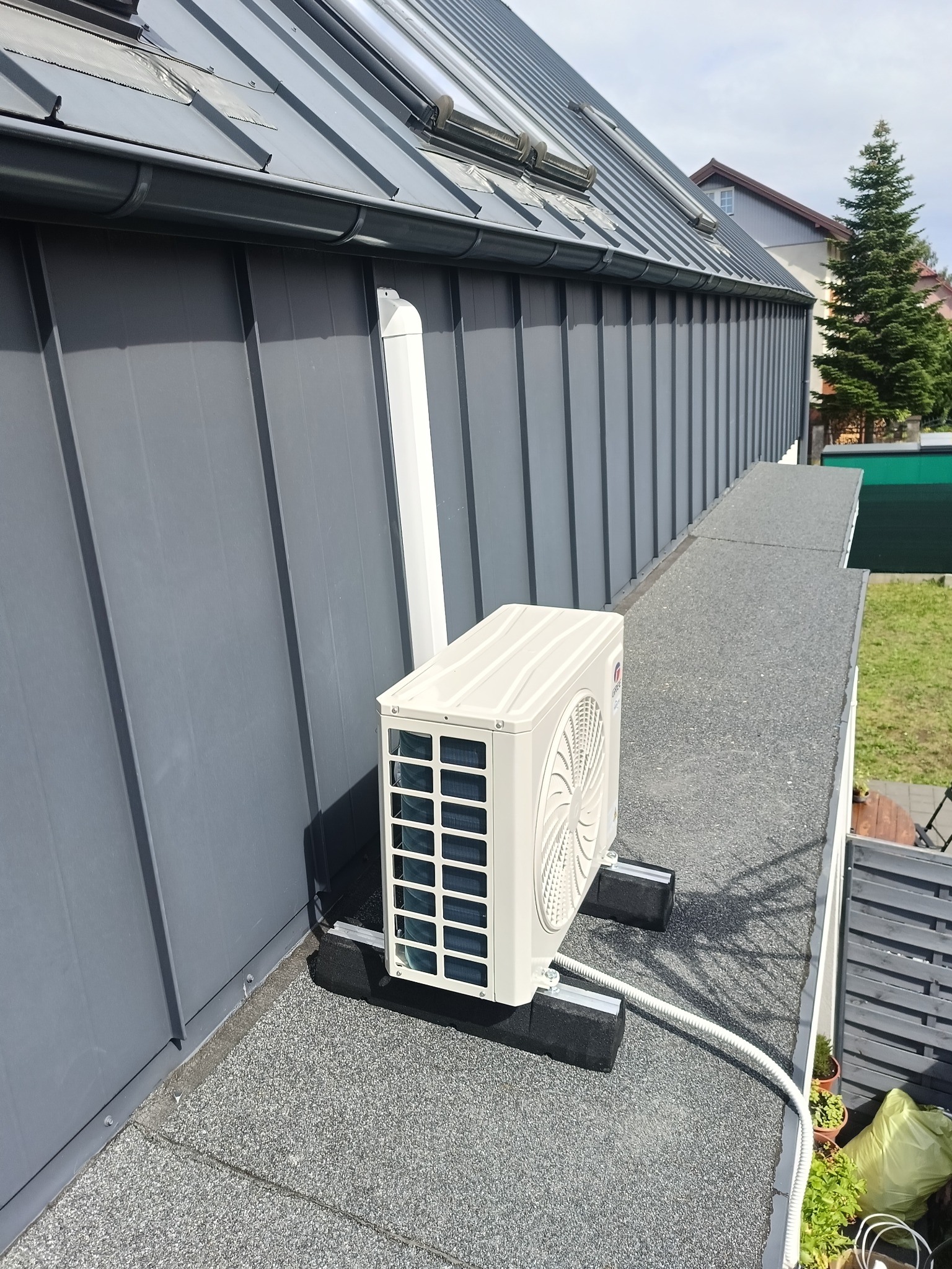 Biała jednostka zewnętrzna klimatyzacji zamontowana na płaskim dachu budynku z szarą blachą na elewacji, widoczne czarne podkładki antywibracyjne i białe rury odprowadzające skropliny.