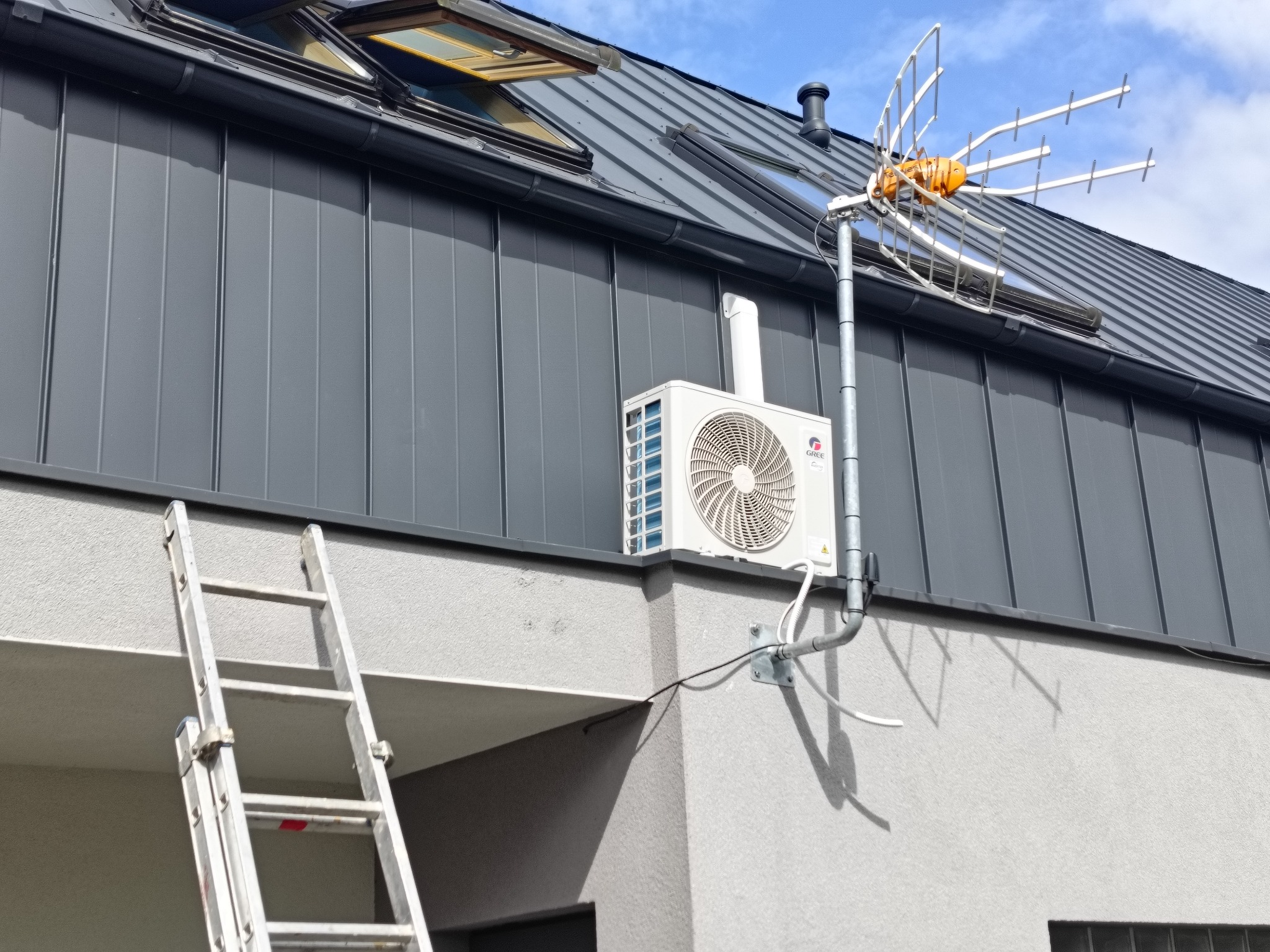 Zamontowana jednostka zewnętrzna klimatyzacji marki Gree na elewacji budynku w Lęborku, widoczna drabina oparta o ścianę, dach z oknami dachowymi w tle.