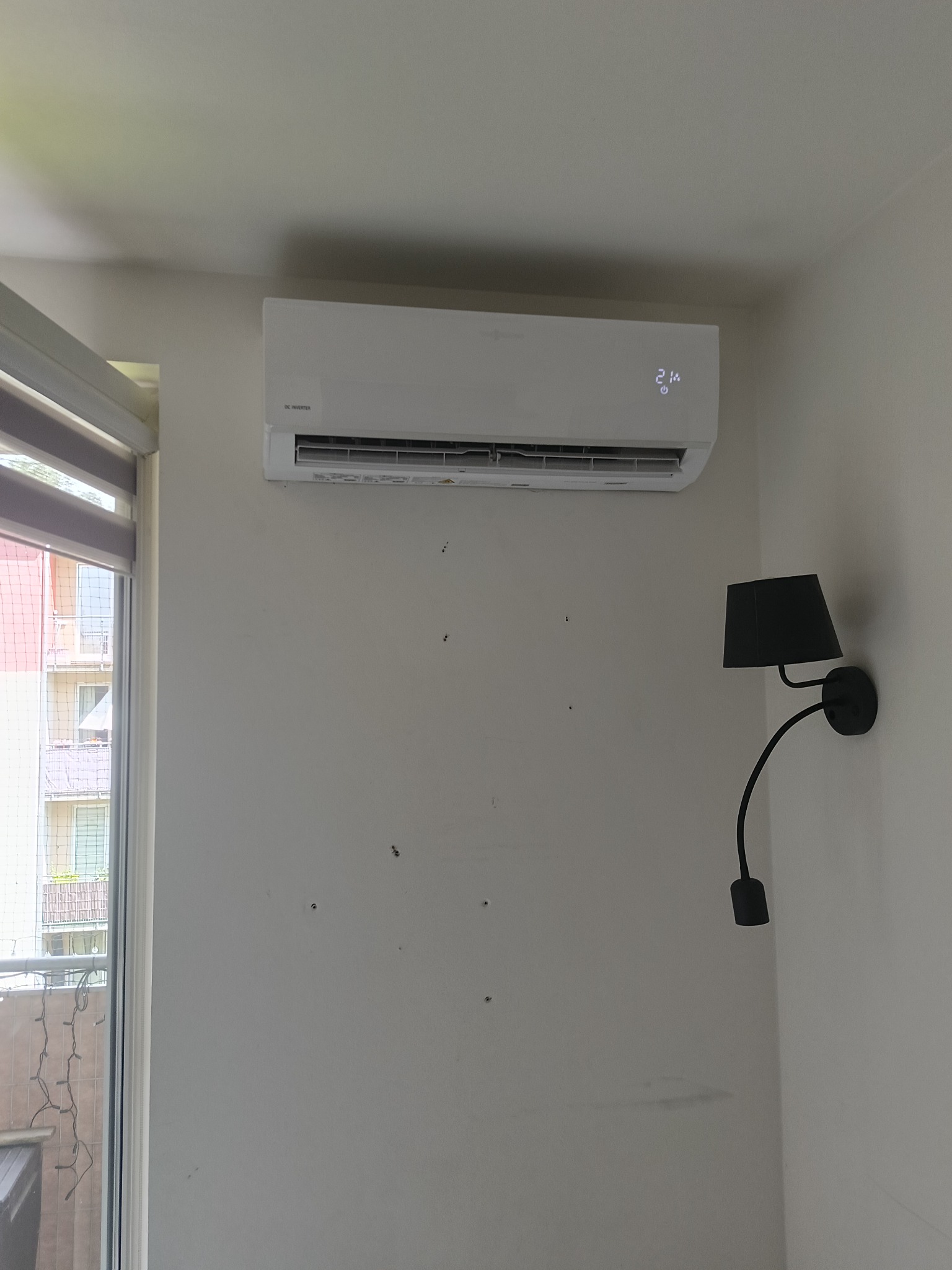 Biały klimatyzator inwerterowy zamontowany na ścianie w jasnym pokoju, widoczne ślady po wcześniejszych mocowaniach, obok czarna lampka ścienna.