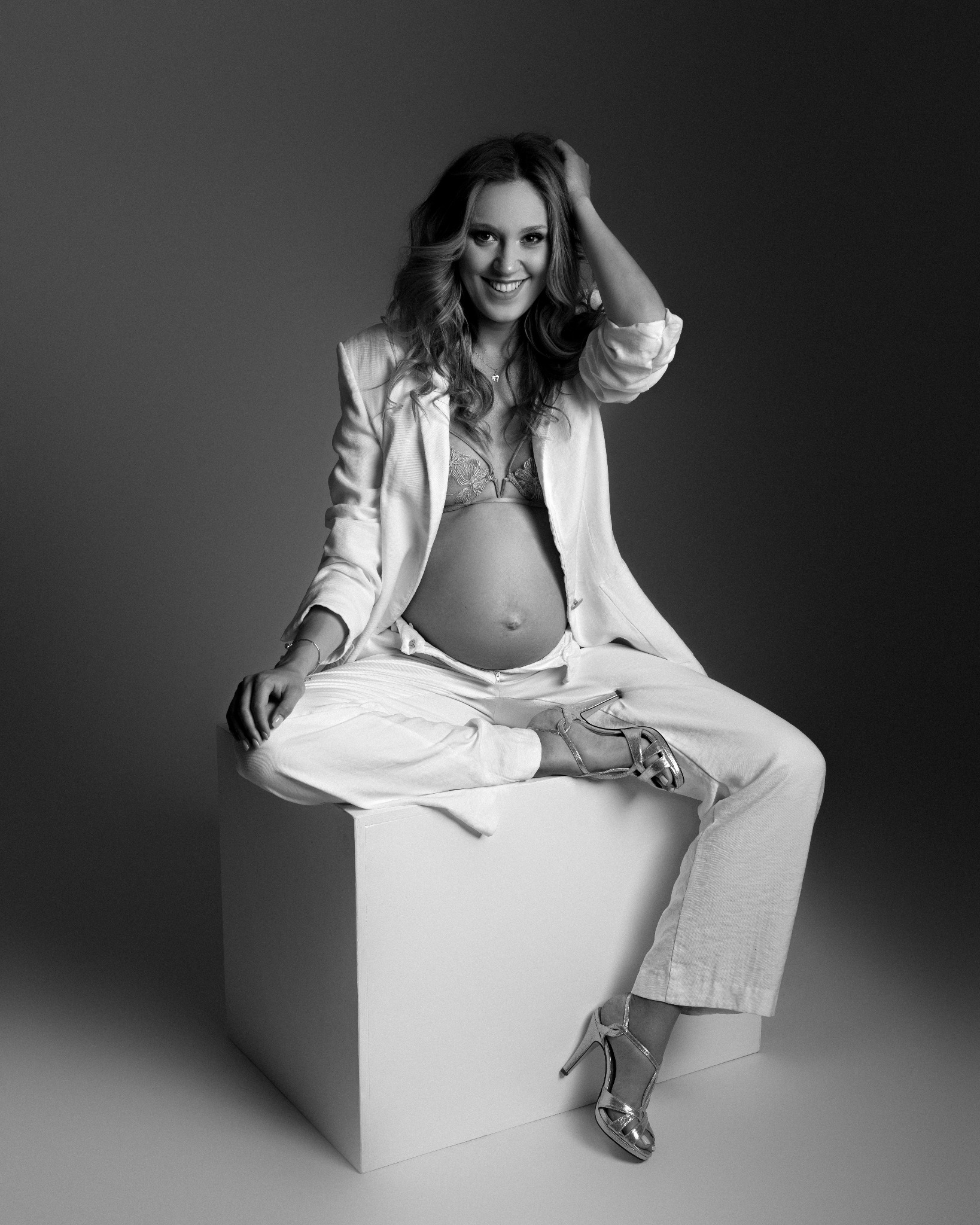 Czarno-białe zdjęcie uśmiechniętej, ciężarnej kobiety siedzącej na białym sześcianie, ubrana w biały garnitur i srebrne szpilki.