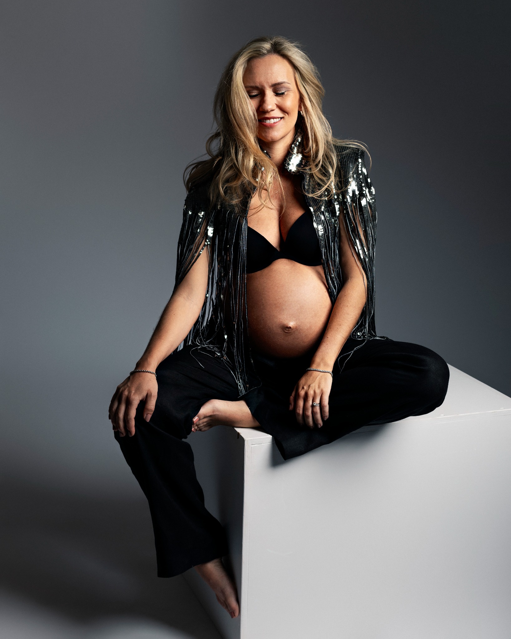 Kobieta w ciąży siedzi na białym sześcianie, ubrana w czarny biustonosz, spodnie i błyszczącą narzutkę z frędzlami, z zamkniętymi oczami.