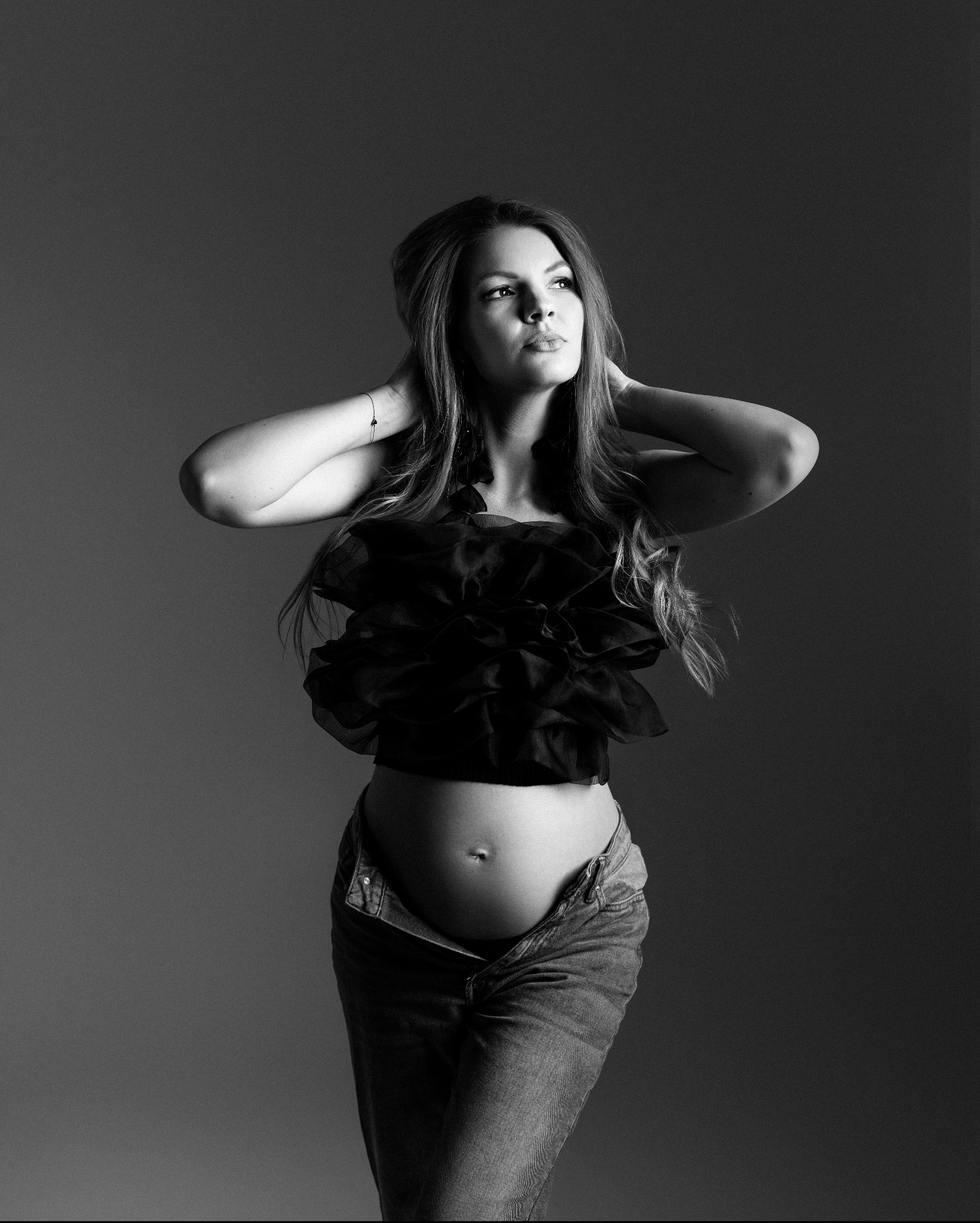 Czarno-białe portretowe ujęcie kobiety w ciąży, pozującej w dżinsach z rozpiętym guzikiem i czarnym topie z falbanami, z rękami uniesionymi za głową i delikatnym uśmiechem, na gładkim szarym tle.