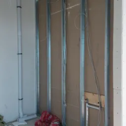 Montaż stelaża z profili stalowych pod płytę karton-gips, widoczne przewody elektryczne, rury kanalizacyjne i izolowane rury grzewcze w surowym wnętrzu.
