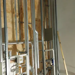 Wnętrze w trakcie prac wykończeniowych: metalowa konstrukcja szkieletowa z izolacją z wełny mineralnej, widoczne rury wentylacyjne i instalacja sanitarna, poziomica oparta o ścianę.