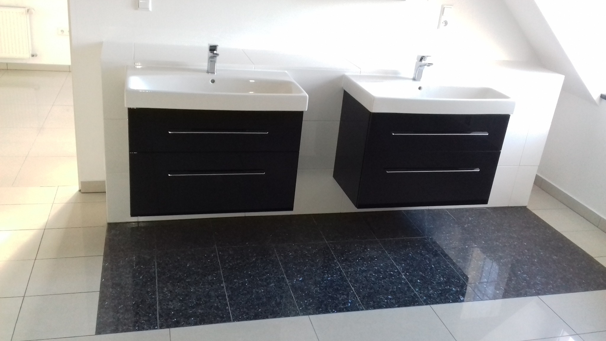 Nowoczesna łazienka z dwoma wiszącymi umywalkami z czarnymi szafkami, chromowanymi bateriami i ciemną, błyszczącą podłogą na tle jasnych płytek.