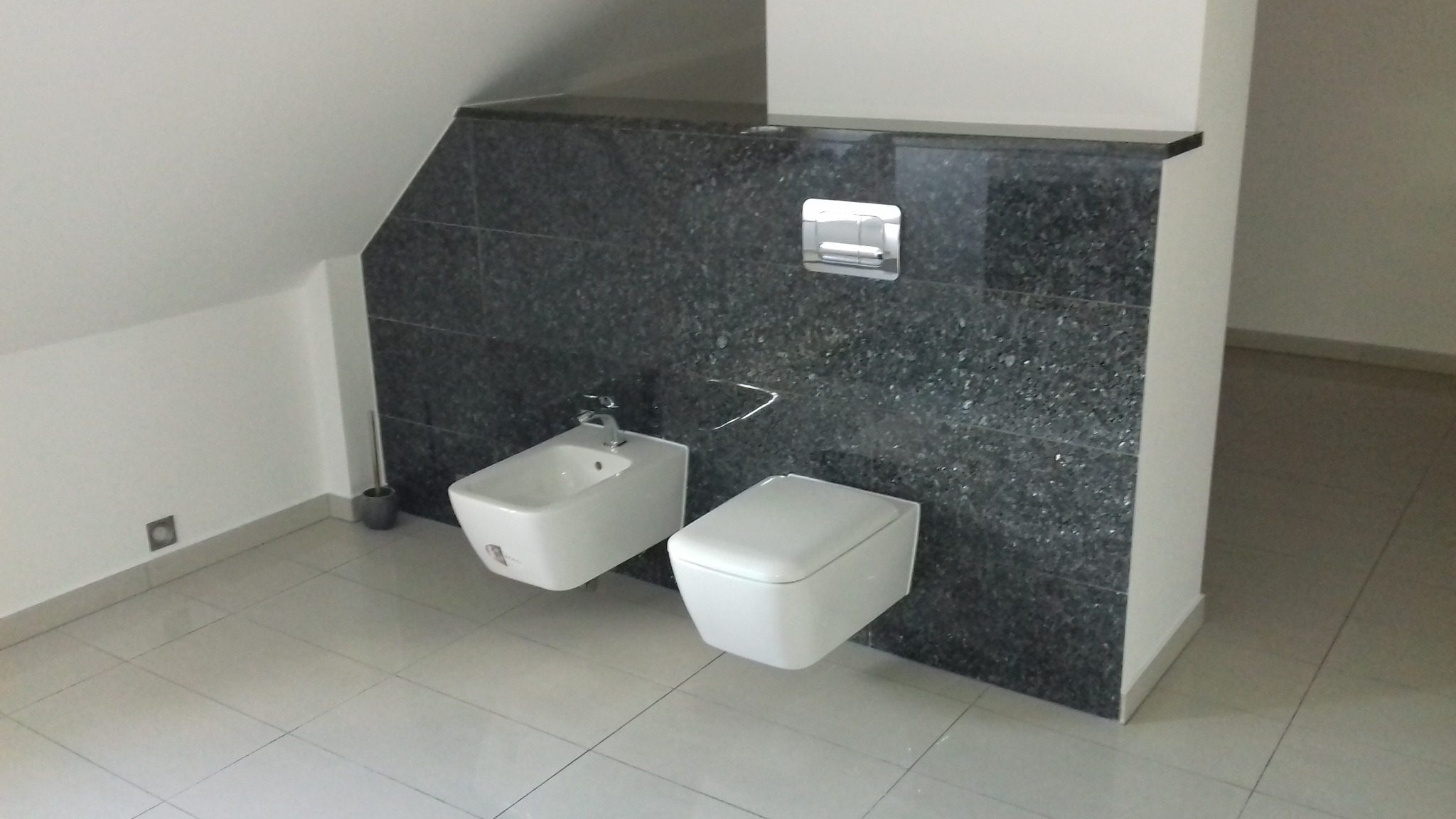 Nowoczesna łazienka na poddaszu: biała ceramika sanitarna (bidet i toaleta podwieszana) na tle ściany wyłożonej ciemnym, błyszczącym kamieniem, z widocznym przyciskiem spłukującym i fragmentem...