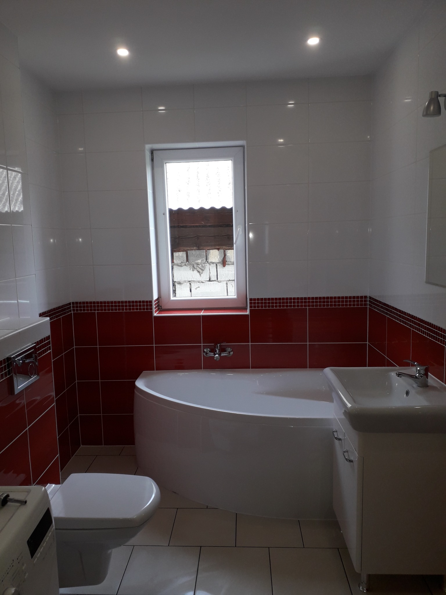 Wykończona łazienka w bieli i czerwieni z narożną wanną, umywalką z szafką i podwieszaną toaletą, oświetlona punktowymi lampami sufitowymi, z oknem z widokiem na mur z białej cegły.