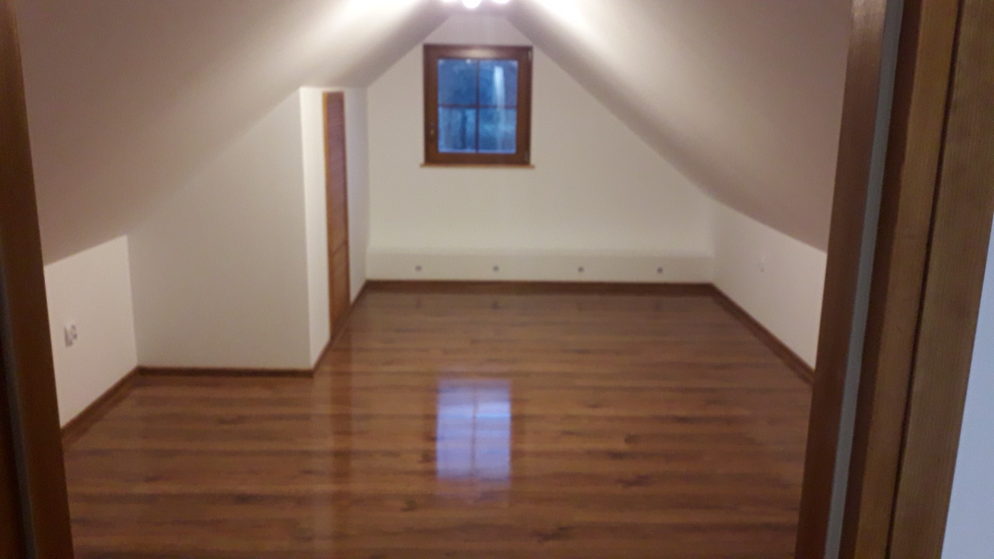 Poddasze z oknem w drewnianej ramie, świeżo wycyklinowana parkietowa podłoga z wyraźnym odbiciem światła, białe ściany i drewniane listwy przypodłogowe, widok zza drewnianej framugi drzwi.