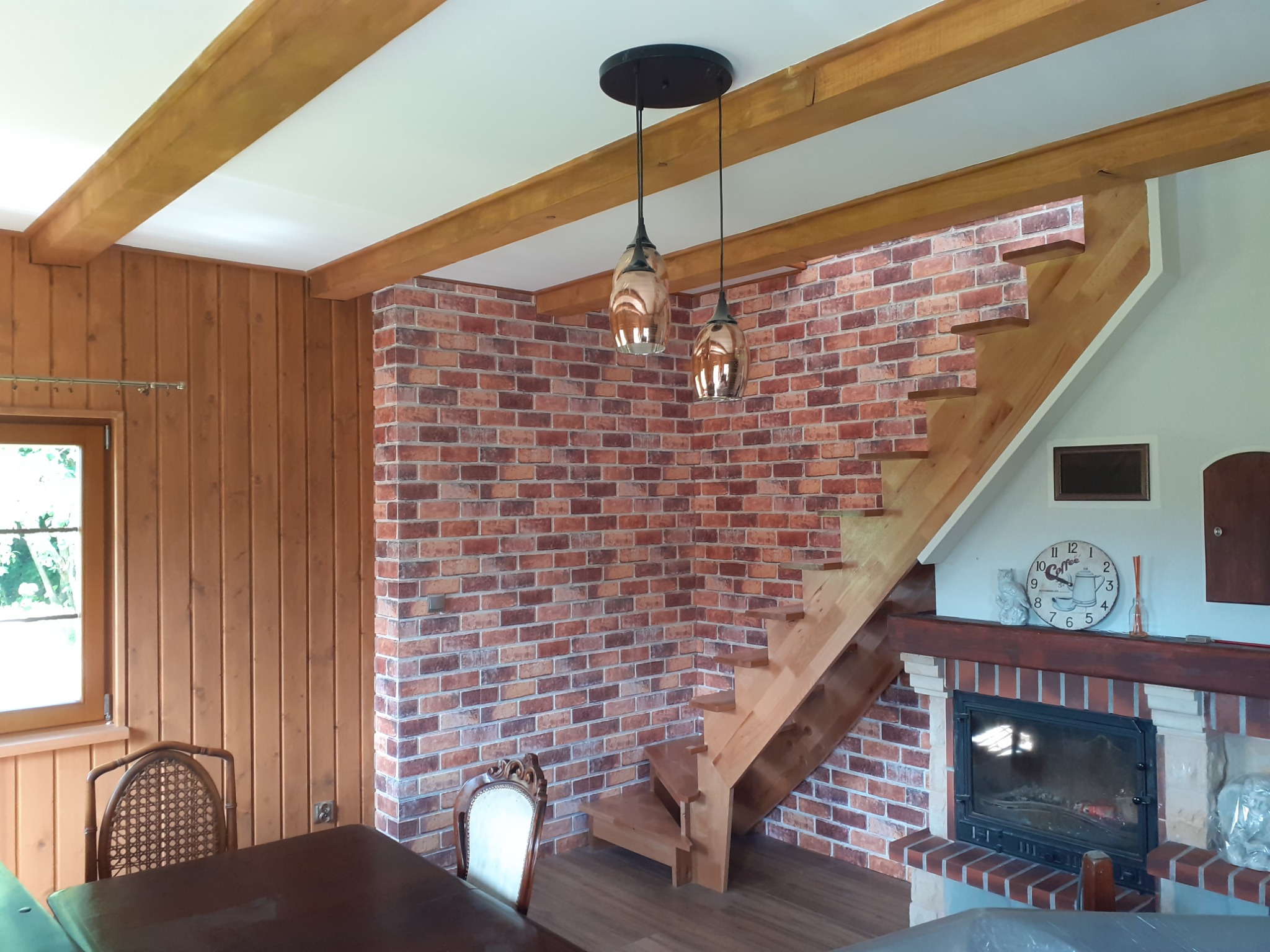 Pokój z ceglaną ścianą, drewnianymi belkami stropowymi, schodami i kominkiem, oświetlony wiszącymi lampami.