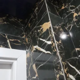Fragment ściany wyłożonej czarnymi płytkami imitującymi marmur z wyraźnymi złotymi i białymi żyłami, widoczna biała futryna drzwi i srebrne listwy wykończeniowe w narożniku.