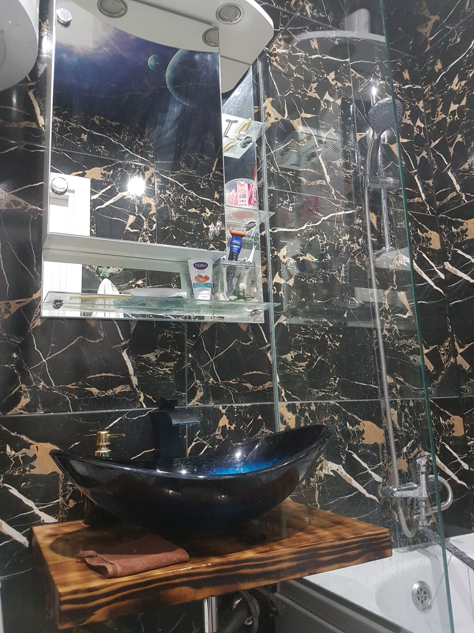 Nowoczesna łazienka z czarnymi płytkami imitującymi marmur, szklaną kabiną prysznicową i umywalką w kształcie łodzi na drewnianym blacie.