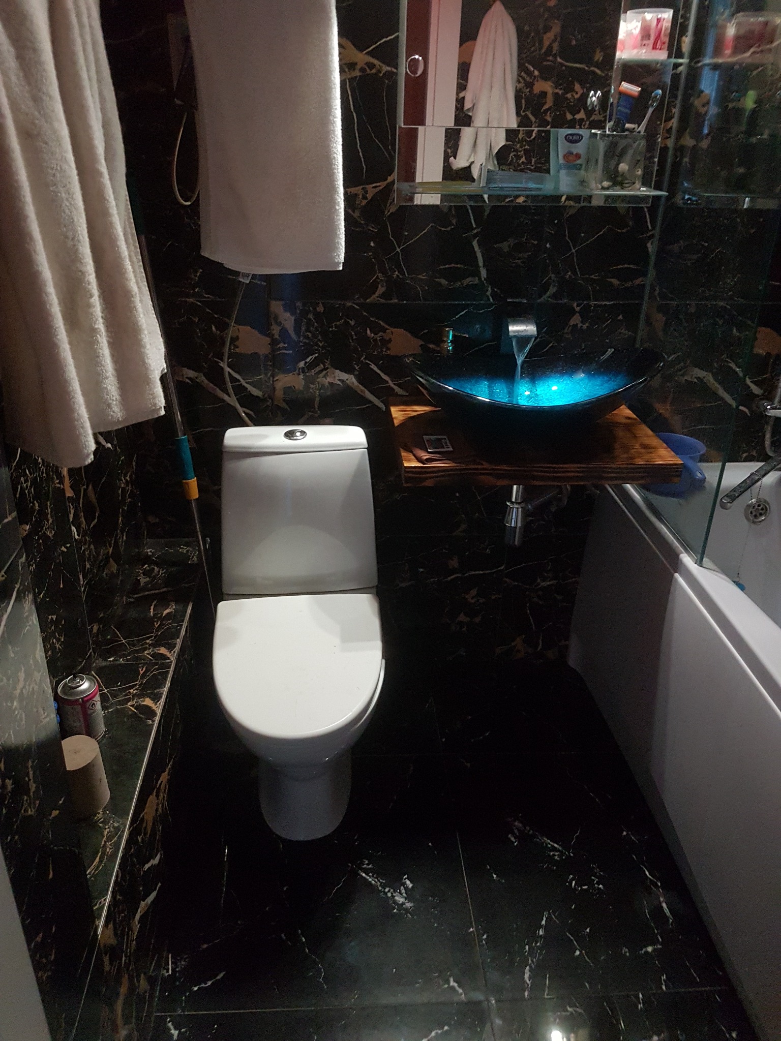 Nowoczesna łazienka z czarnymi marmurowymi płytkami, podświetlaną umywalką z niebieskim światłem, białą toaletą i ręcznikami.