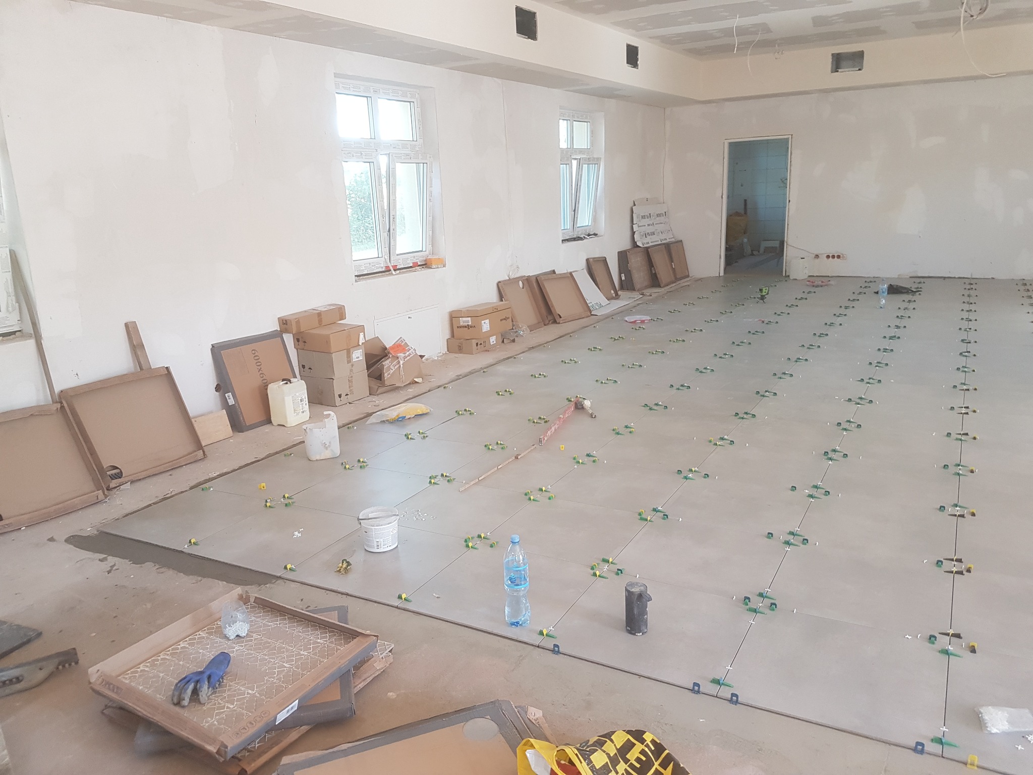 Proces układania szarych płytek podłogowych w dużym, surowym pomieszczeniu z oknami i otwartymi kartonami płytek. Widoczne krzyżyki dystansowe i poziomica laserowa.