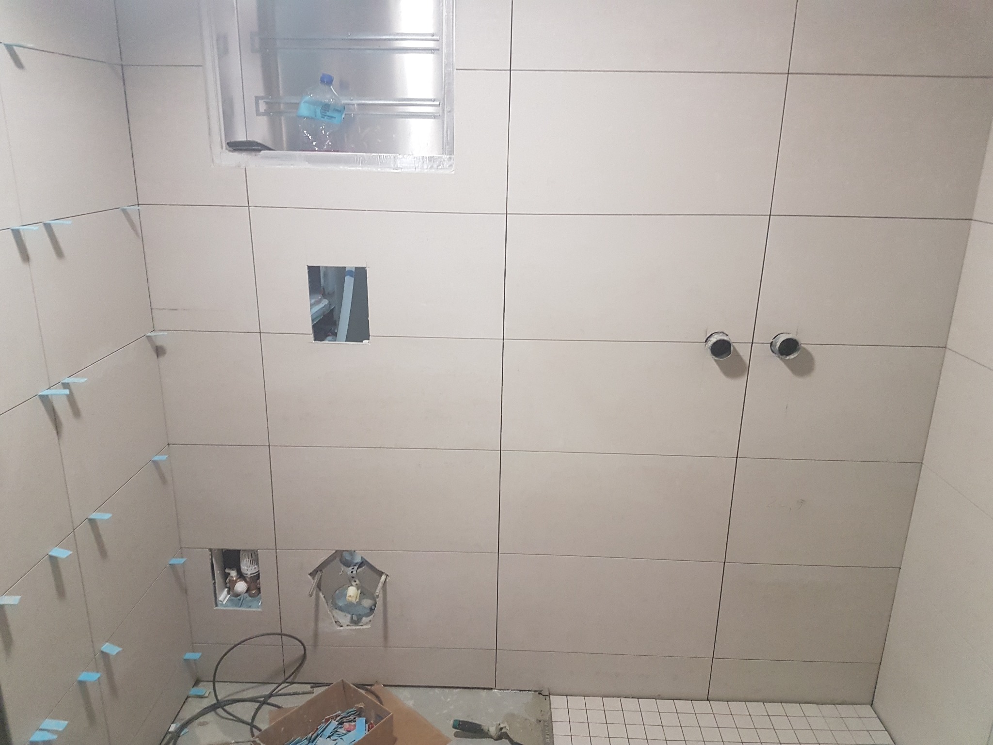 Łazienka w trakcie wykańczania: beżowe płytki ceramiczne ułożone na ścianach z niebieskimi krzyżykami dystansowymi, widoczne otwory na instalacje, pudełko z akcesoriami i kielnia na podłodze...