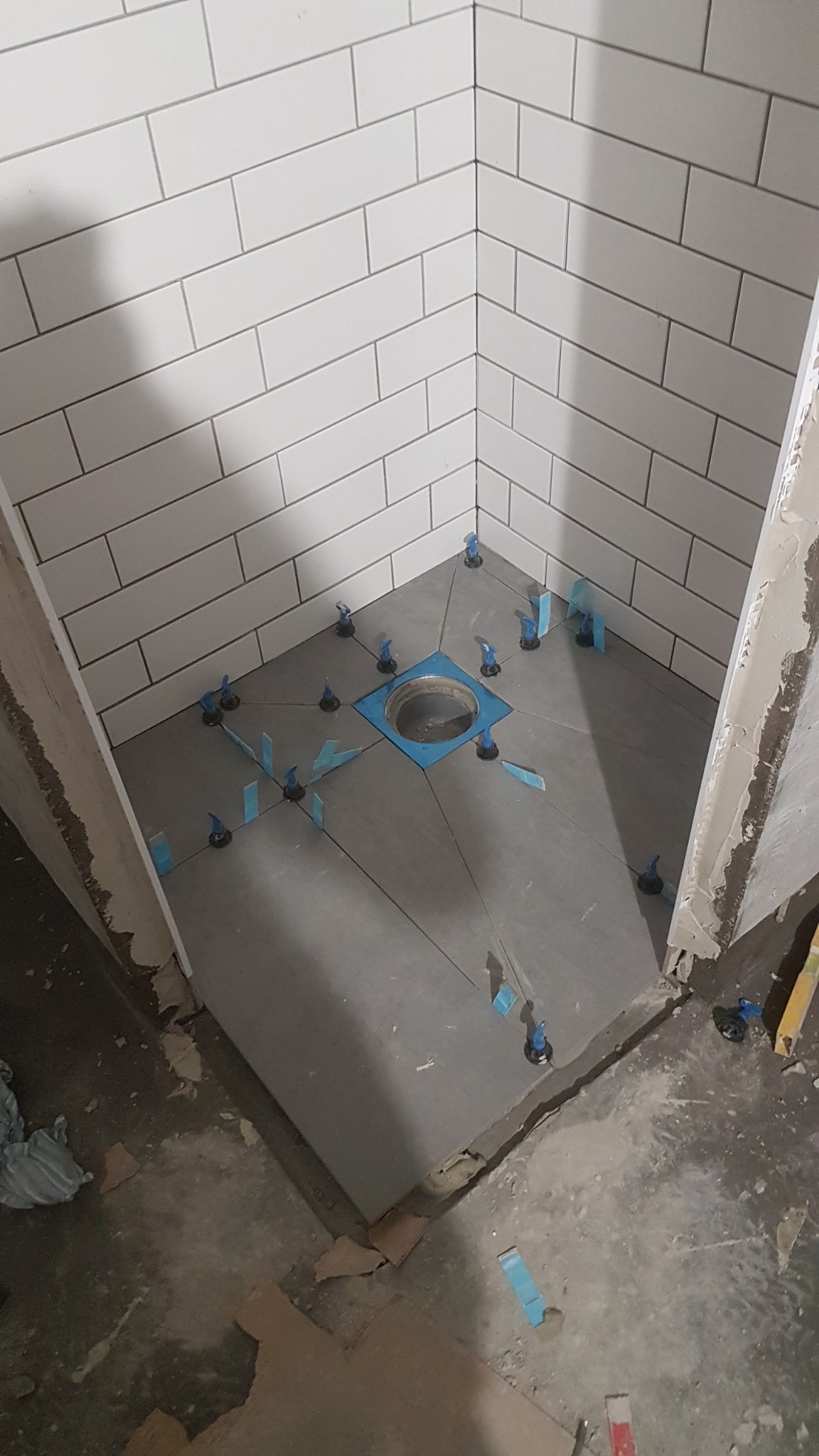 Prysznic w trakcie remontu: ułożone szare płytki podłogowe ze stabilizatorami i niebieskimi klinami wokół odpływu, białe prostokątne płytki na ścianach, widoczne ślady prac remontowych.