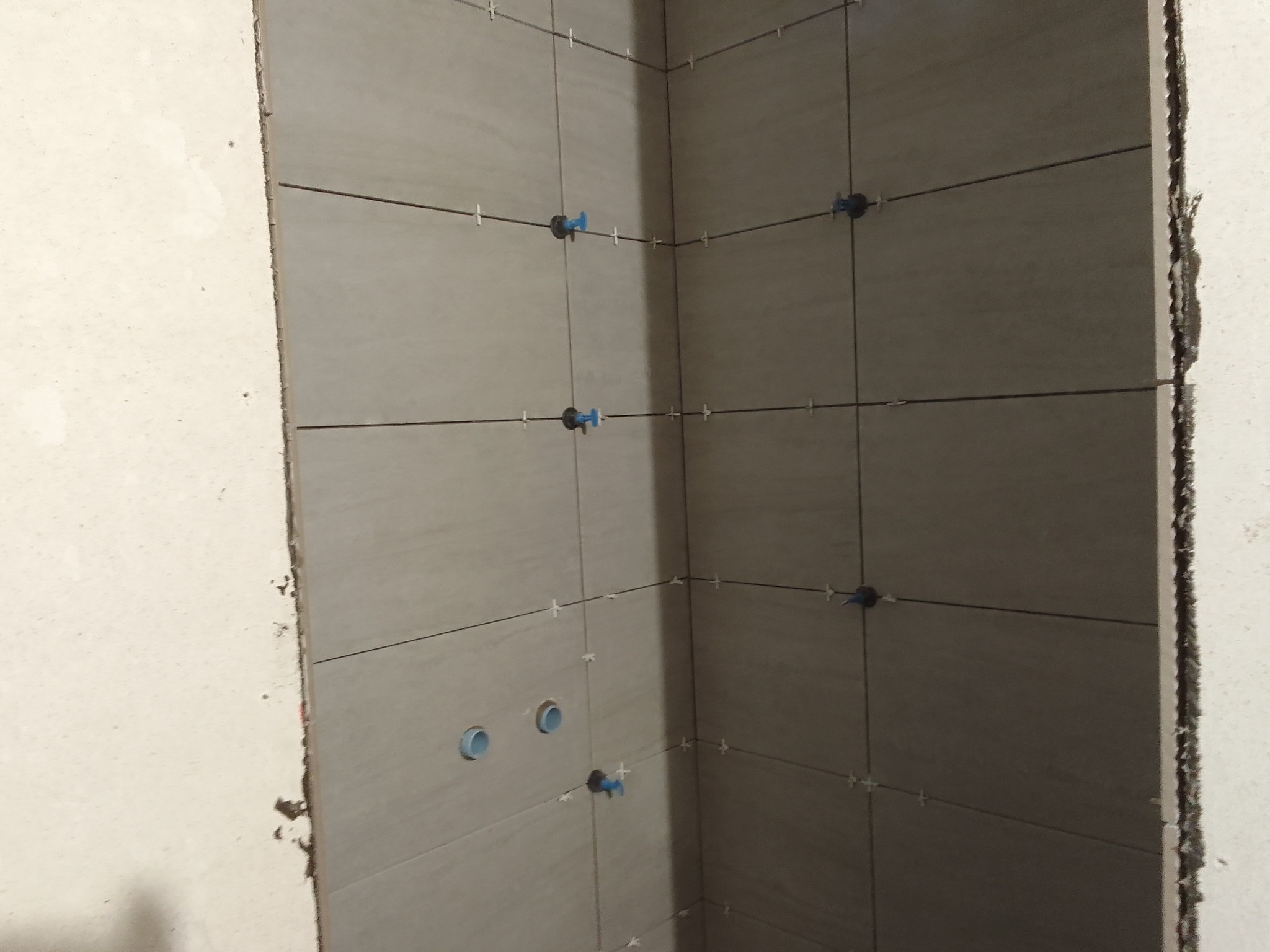 Ściana w trakcie układania płytek ceramicznych w narożniku, widoczne krzyżyki dystansowe i otwory na rury.