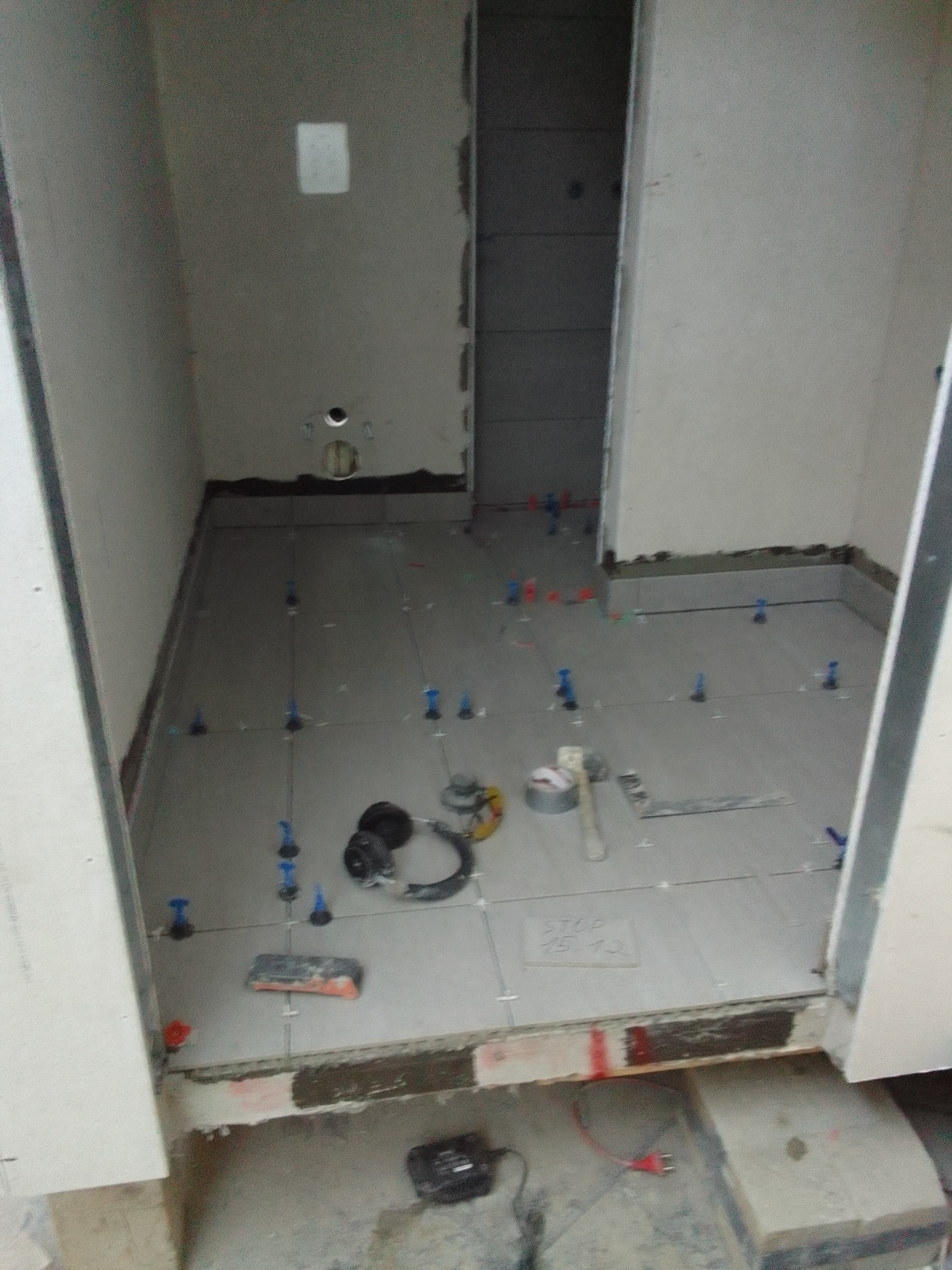 Wnętrze łazienki w trakcie remontu: ułożone płytki podłogowe z niebieskimi krzyżykami dystansowymi, widoczne narzędzia glazurnicze i niedokończone ściany.