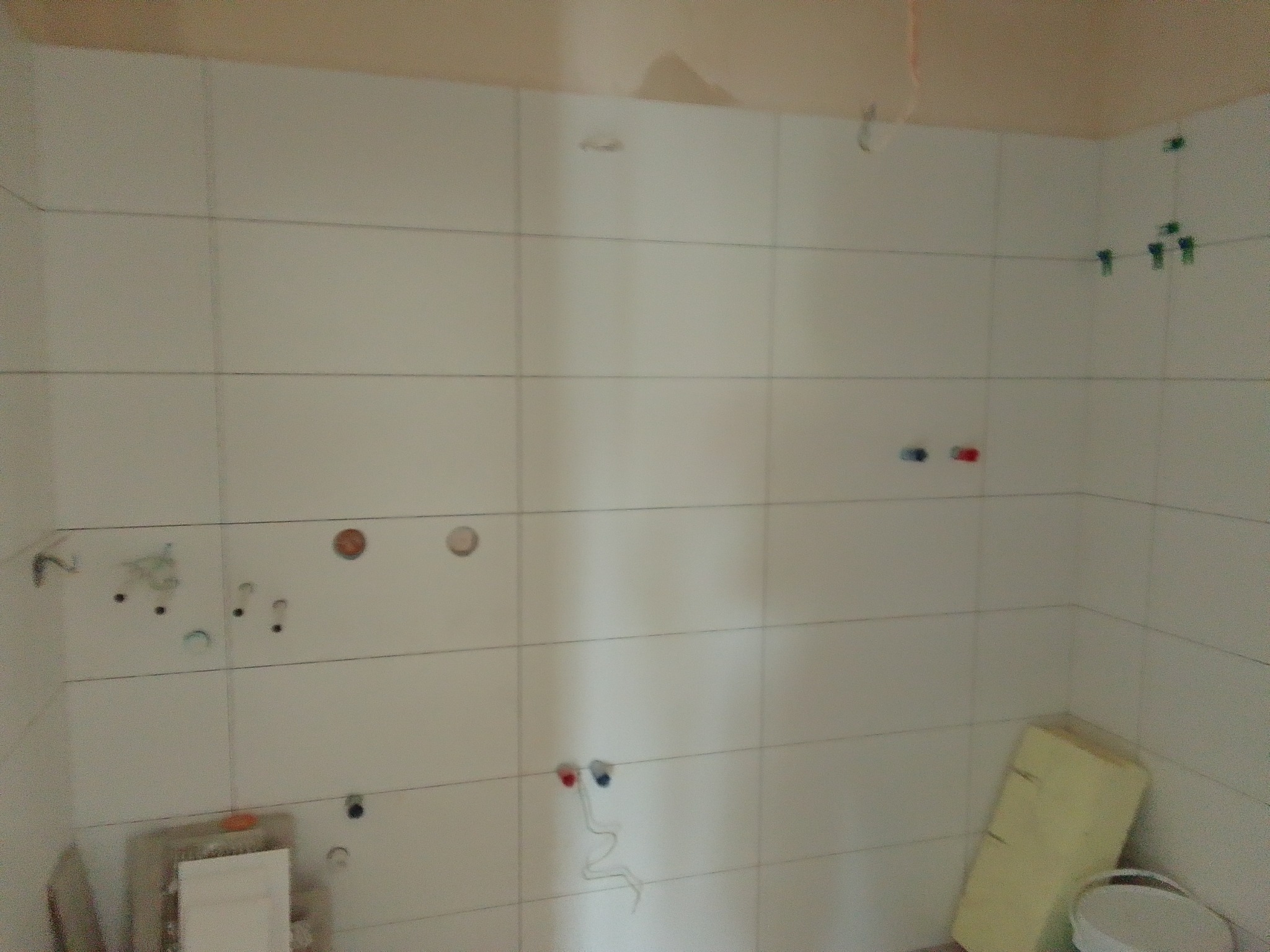 Ściana łazienki w trakcie układania białych płytek, widoczne otwory na instalacje, poziomice i plastikowe krzyżyki dystansowe.