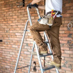 Elektryk na drabinie, z narzędziami przy pasie, pracuje przy instalacji elektrycznej w surowym wnętrzu z ceglaną ścianą.