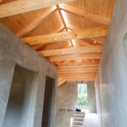 Surowy stan domu: betonowe ściany, drewniana więźba dachowa, schody. Widok wnętrza z otworami drzwiowymi i oknem na zewnątrz. Etap budowy domu.