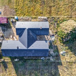 Wymarzone Domy - Zdjęcie z drona ukończonego domu z ciemną dachówką, widoczne ułożone drewno, czerwony blaszak i niezagospodarowany teren. Kąt widzenia z góry, w słoneczny dzień.