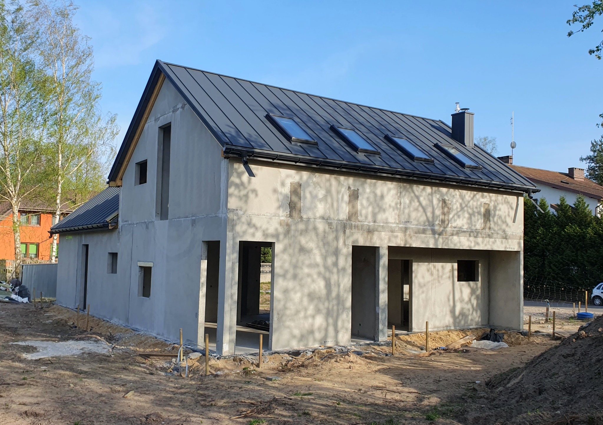 Surowy, dwukondygnacyjny dom z szarą blachodachówką i oknami dachowymi, w trakcie budowy, z widocznymi otworami okiennymi i drzwiowymi, otoczony ziemią.