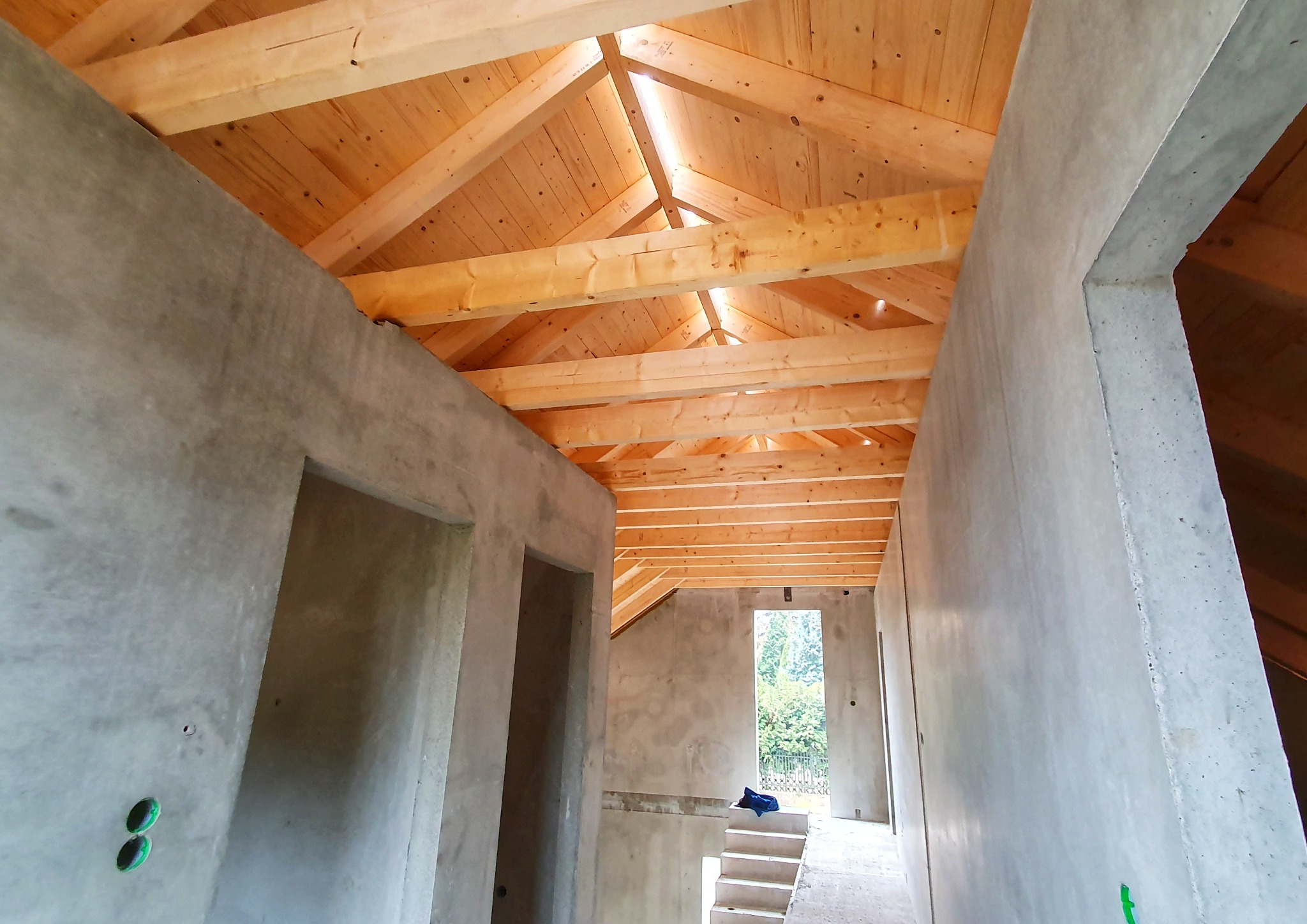 Surowy stan domu: betonowe ściany, drewniana więźba dachowa, schody. Widok wnętrza z otworami drzwiowymi i oknem na zewnątrz. Etap budowy domu.