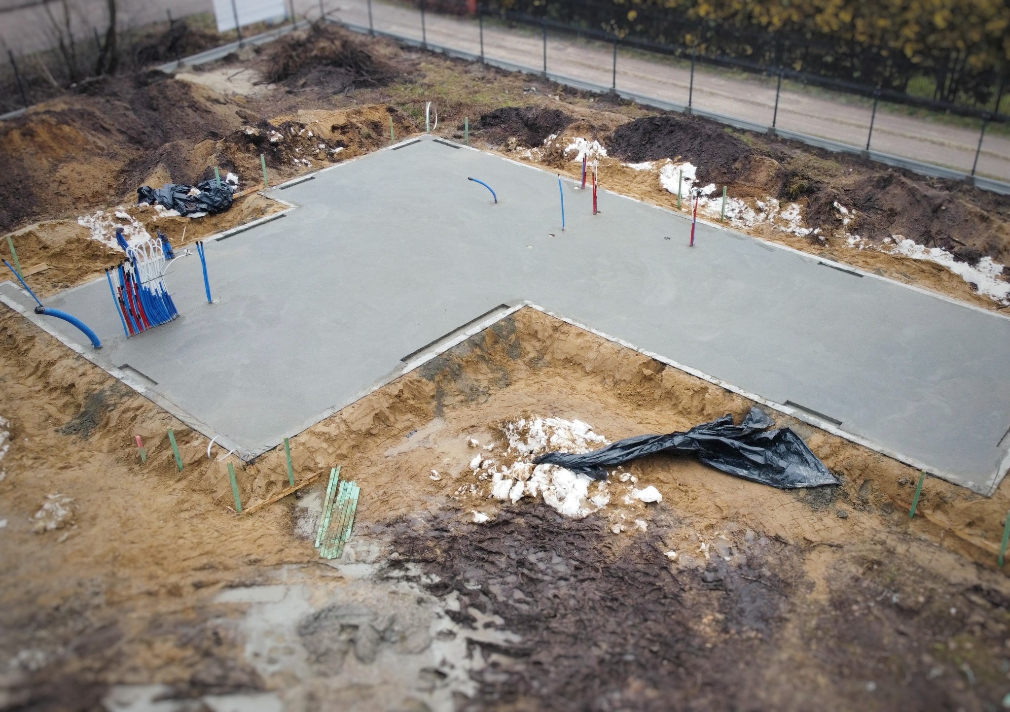 Wylany fundament w kształcie litery L, z wystającymi rurami instalacyjnymi w kolorach niebieskim i czerwonym. Widoczne szalunki i przygotowany teren wokół fundamentu.