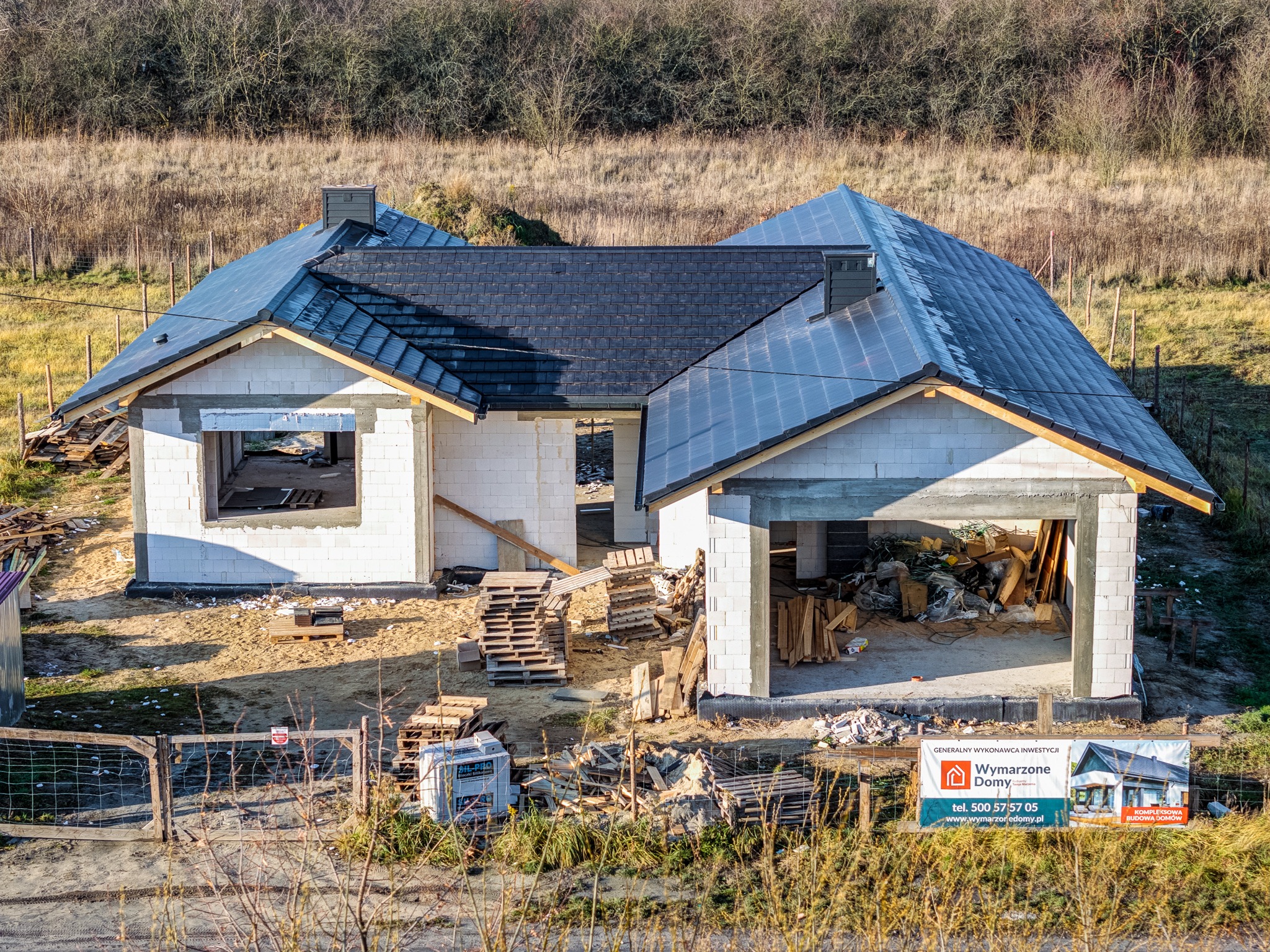 Wznoszony dom murowany z dachem z ciemnej dachówki, widoczny garaż i białe bloczki. Na pierwszym planie ogrodzenie i reklama firmy budowlanej.