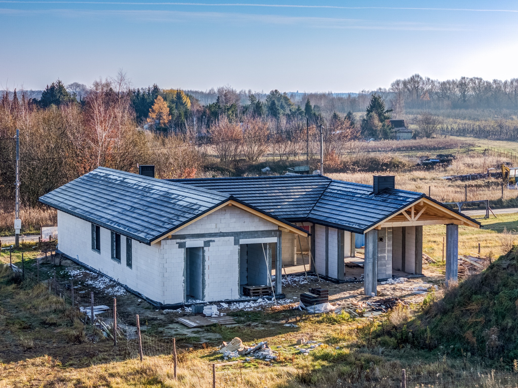 Wznoszenie domu: surowy stan budynku z białych bloczków, widoczne zadaszenie i komin. Otoczenie to trawa i drzewa, co sugeruje lokalizację na obrzeżach miasta.
