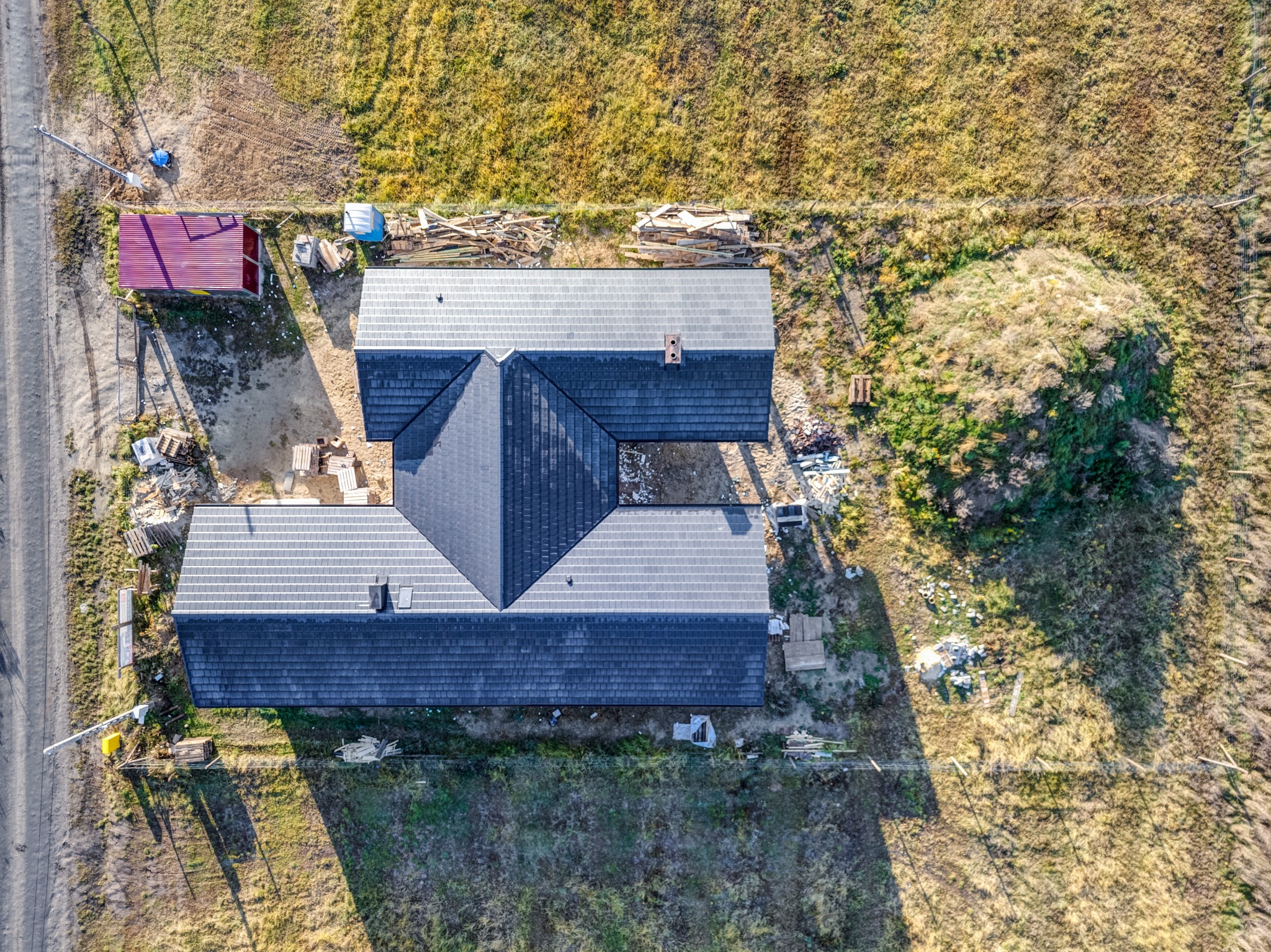 Zdjęcie z drona ukończonego domu z ciemną dachówką, widoczne ułożone drewno, czerwony blaszak i niezagospodarowany teren. Kąt widzenia z góry, w słoneczny dzień.