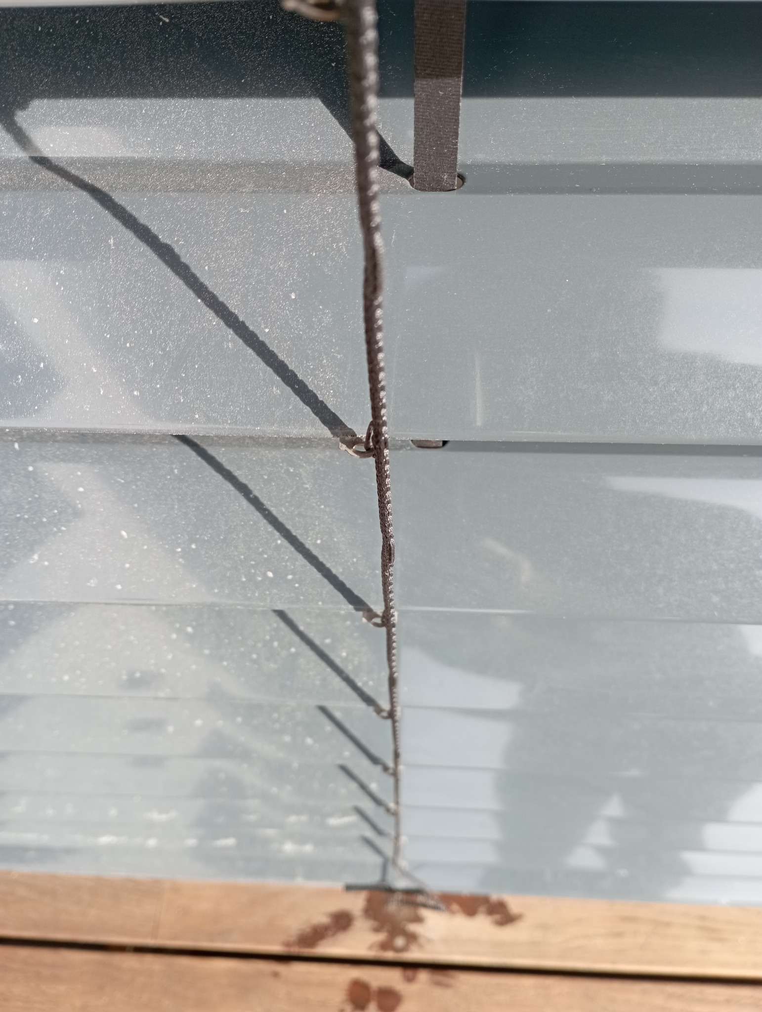 Zabrudzone, szare żaluzje poziome widziane z dołu, z widocznymi smugami i zaciekami na drewnianym parapecie, oświetlone jasnym, naturalnym światłem.