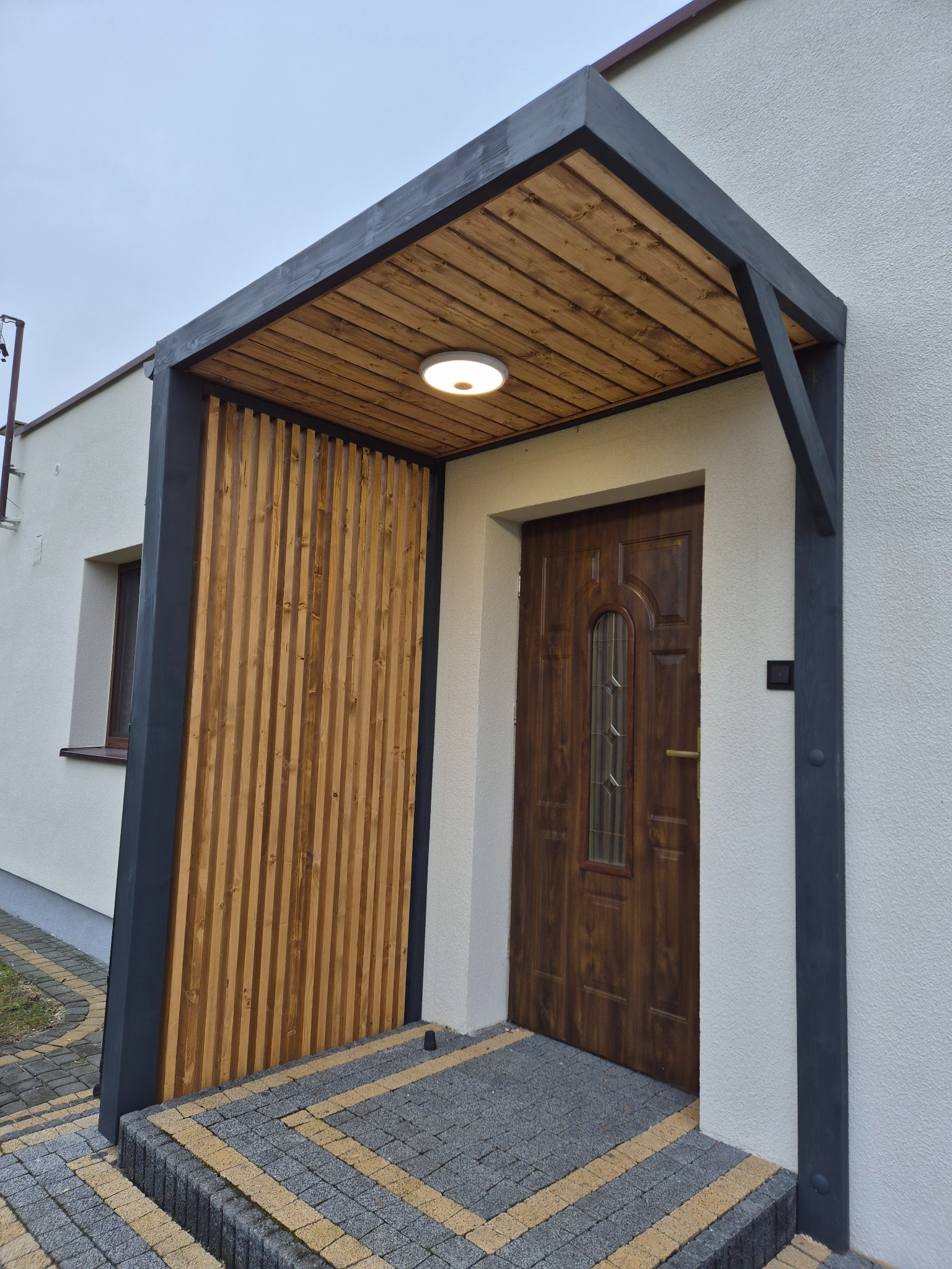 Zadaszenie wejścia z drewnianą boazerią i oświetleniem, wsparte na ciemnych, metalowych słupach. Schody wejściowe wyłożone kostką brukową w dwóch kolorach.
