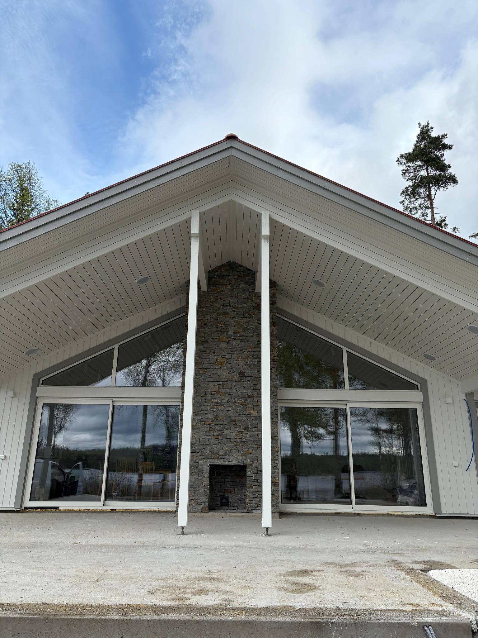 Nowoczesny dom z dużymi oknami i kamiennym kominem. Widoczne elementy drewniane w konstrukcji dachu i wykończeniu elewacji. Minimalistyczny styl architektoniczny.