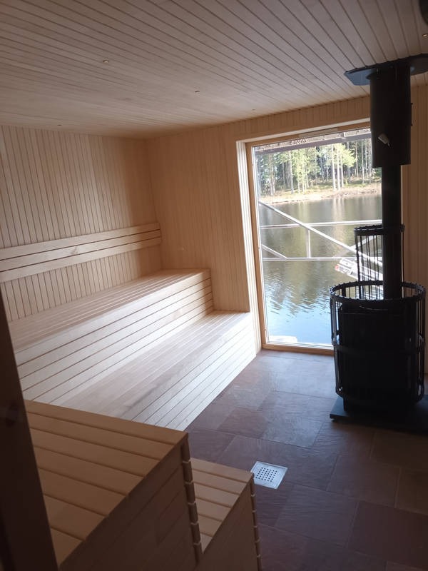 Jasne, drewniane wnętrze sauny z piecem i widokiem na jezioro przez okno. Drewniane ławy i wykończenie ścian tworzą relaksującą atmosferę.
