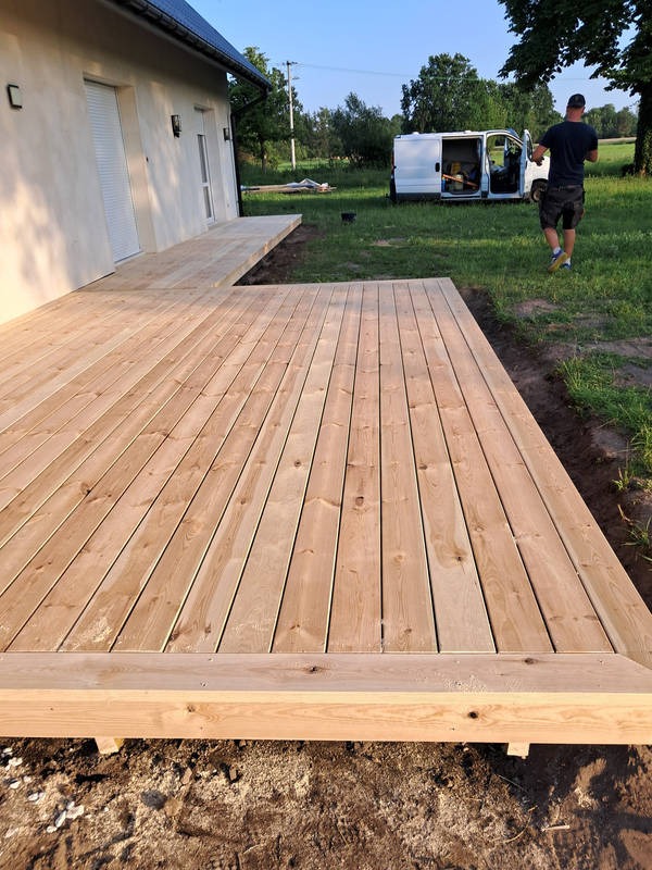 Nowy taras z jasnego drewna przy domu z białymi roletami, z widocznym fragmentem ogrodu i samochodem dostawczym w tle, w słoneczny dzień.