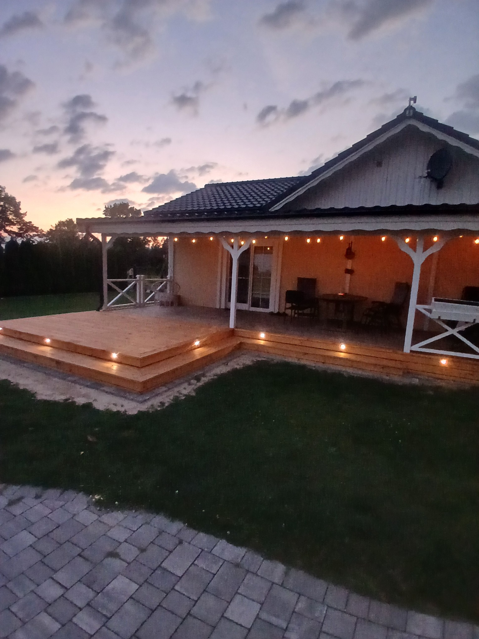 Oświetlony drewniany taras z niskimi schodami prowadzącymi do domu, widok o zmierzchu. Lampki punktowe w podłodze tarasu i girlandy świetlne pod dachem.