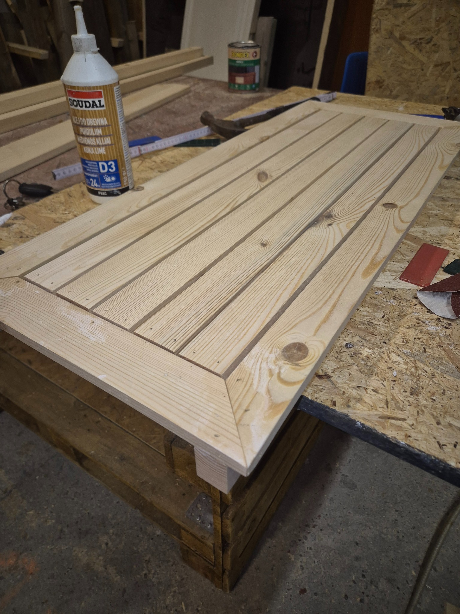 Surowy, drewniany blat z listew, z ramą, na warsztacie. Widoczny klej do drewna, miarka i materiały ścierne. Blat ułożony na drewnianej konstrukcji.