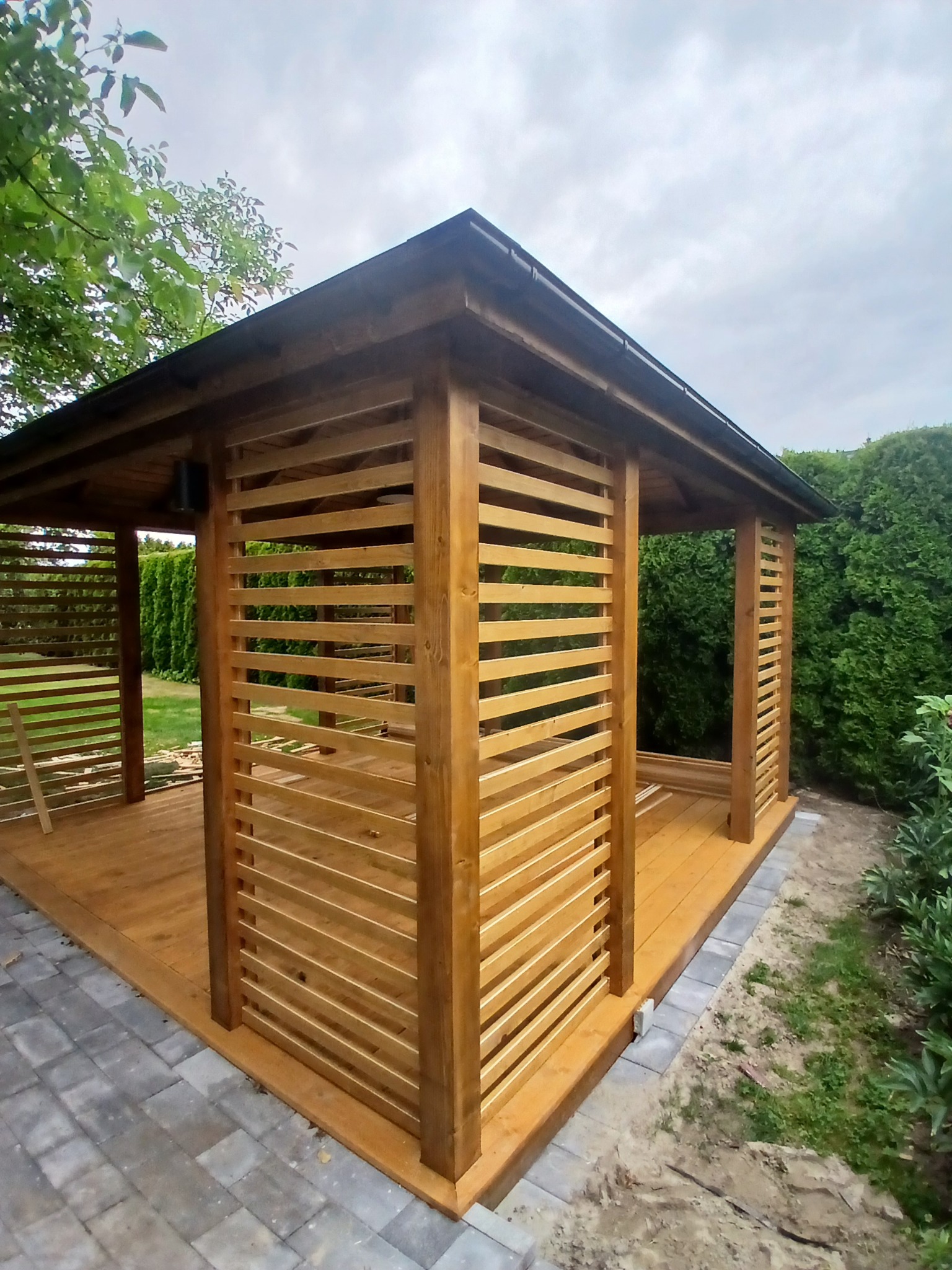 Drewniana altana z poziomymi listwami, widok z zewnątrz, na tle zieleni. Świeżo zaimpregnowane drewno, regularne odstępy między listwami.