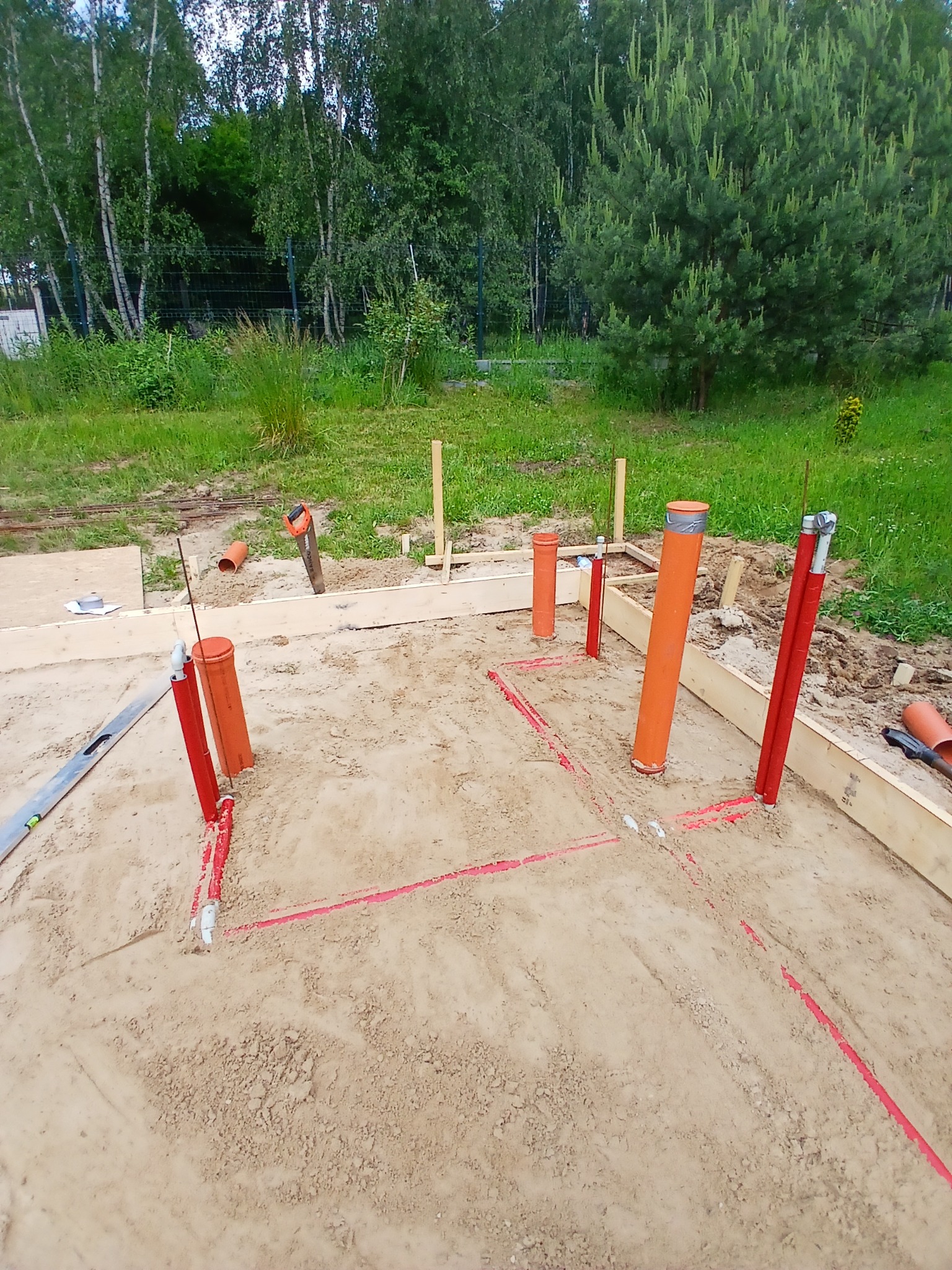 Fundament z instalacją: czerwone rury i pomarańczowe piony w piasku, drewniana rama. Widok z góry na plac budowy z zielenią w tle.