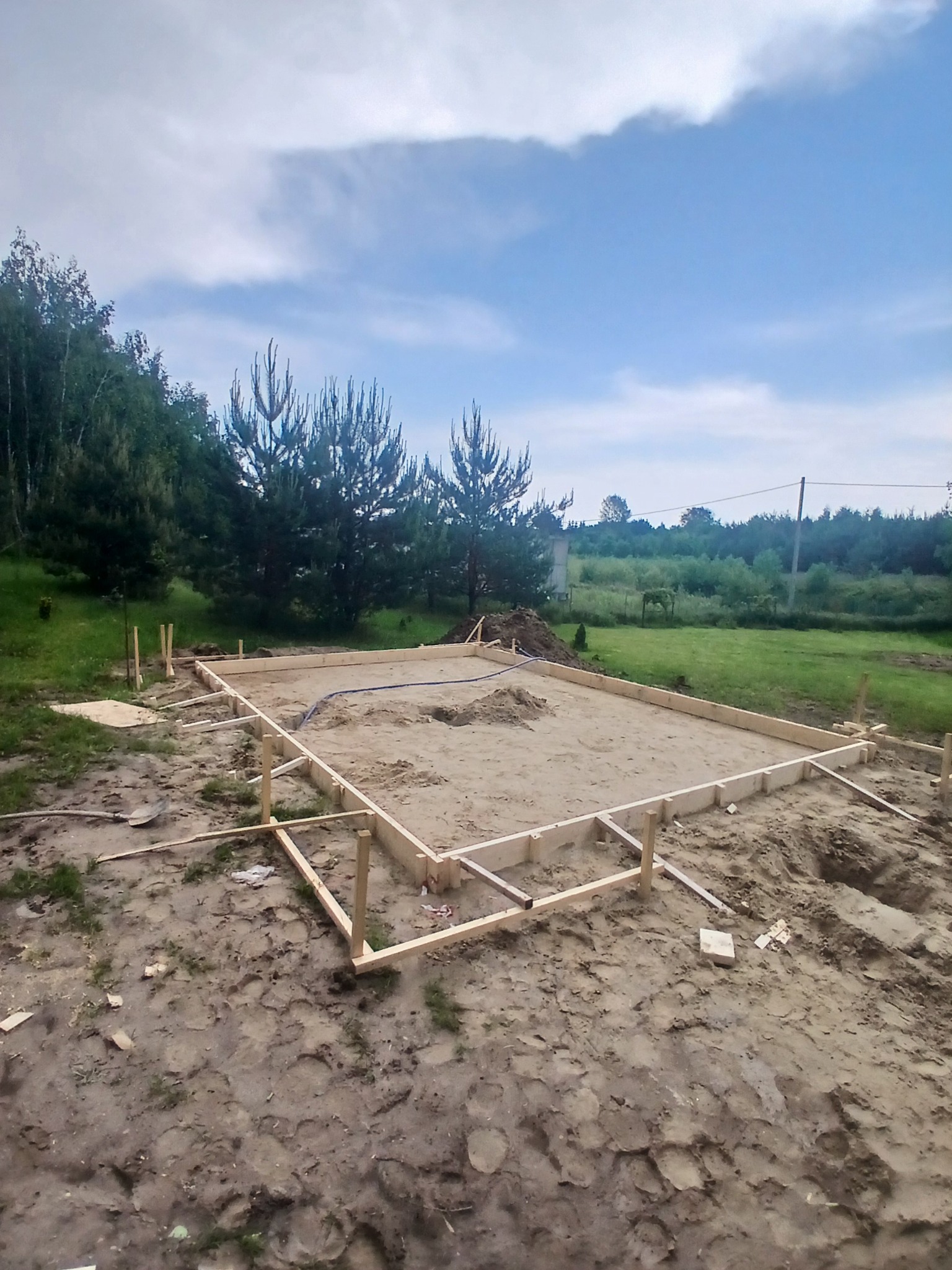 Przygotowanie terenu pod budowę: drewniany szalunek fundamentów na piaszczystej działce, widoczne drzewa w tle, błękitne niebo z chmurami. Etap wylewania fundamentów.