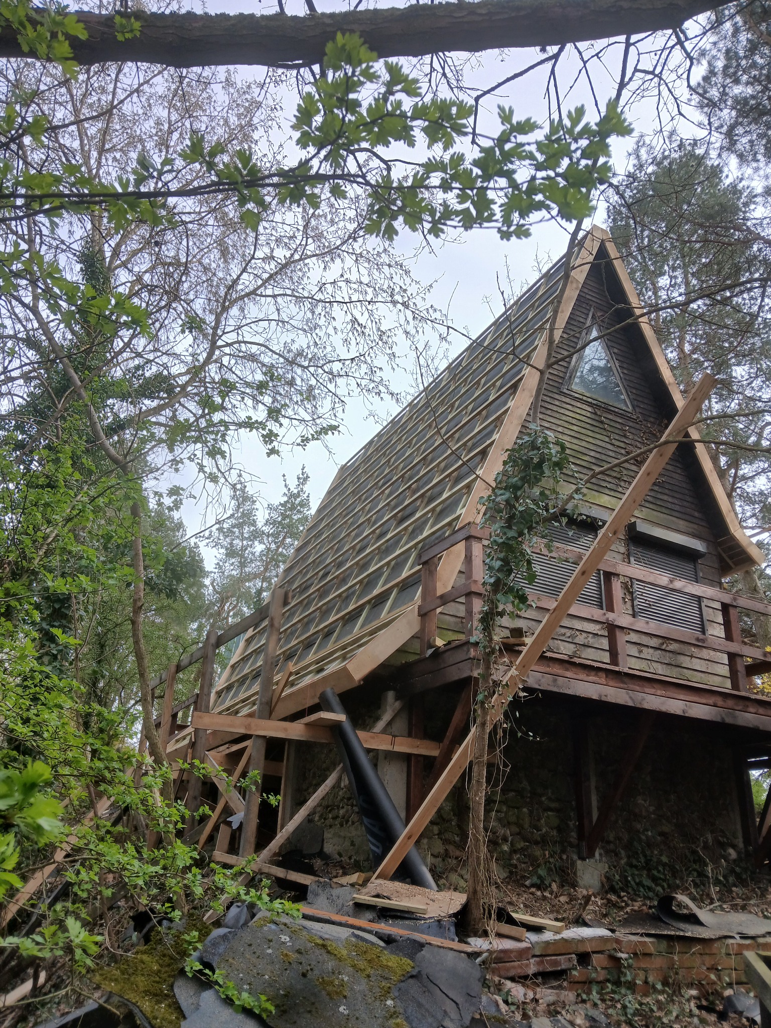 Nietypowa konstrukcja drewniana w lesie, przypominająca domek letniskowy w trakcie budowy. Widoczne elementy szkieletu dachu i drewniane wykończenia. Otoczenie leśne.